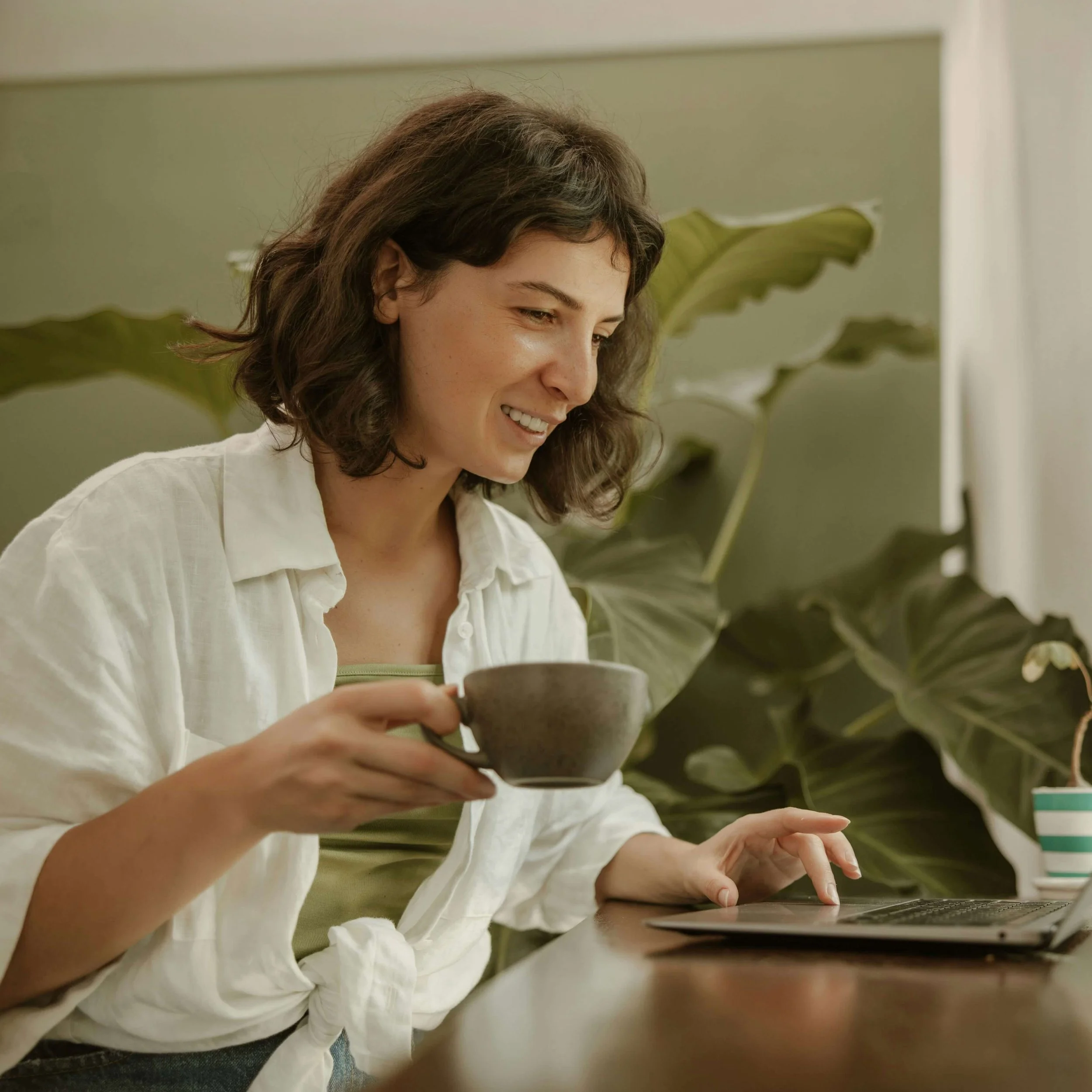 A woman with curly brown hair wearing a white shirt sitting at a table, holding a coffee cup, with a large green houseplant in the background.