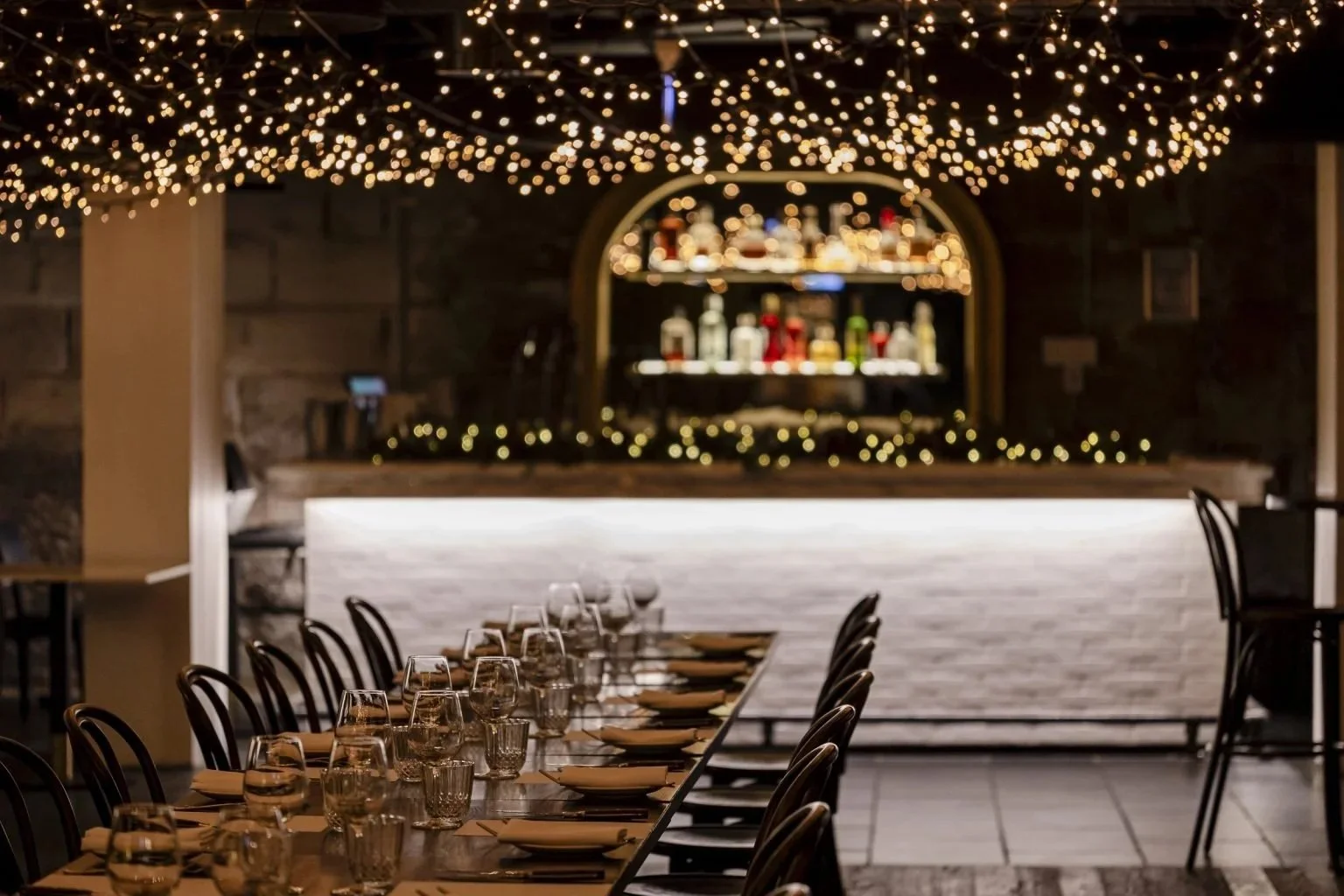 Elegant restaurant table set for dinner with plates, glasses, and napkins, surrounded by beige quilted chairs, and a wine cellar wall in the background.