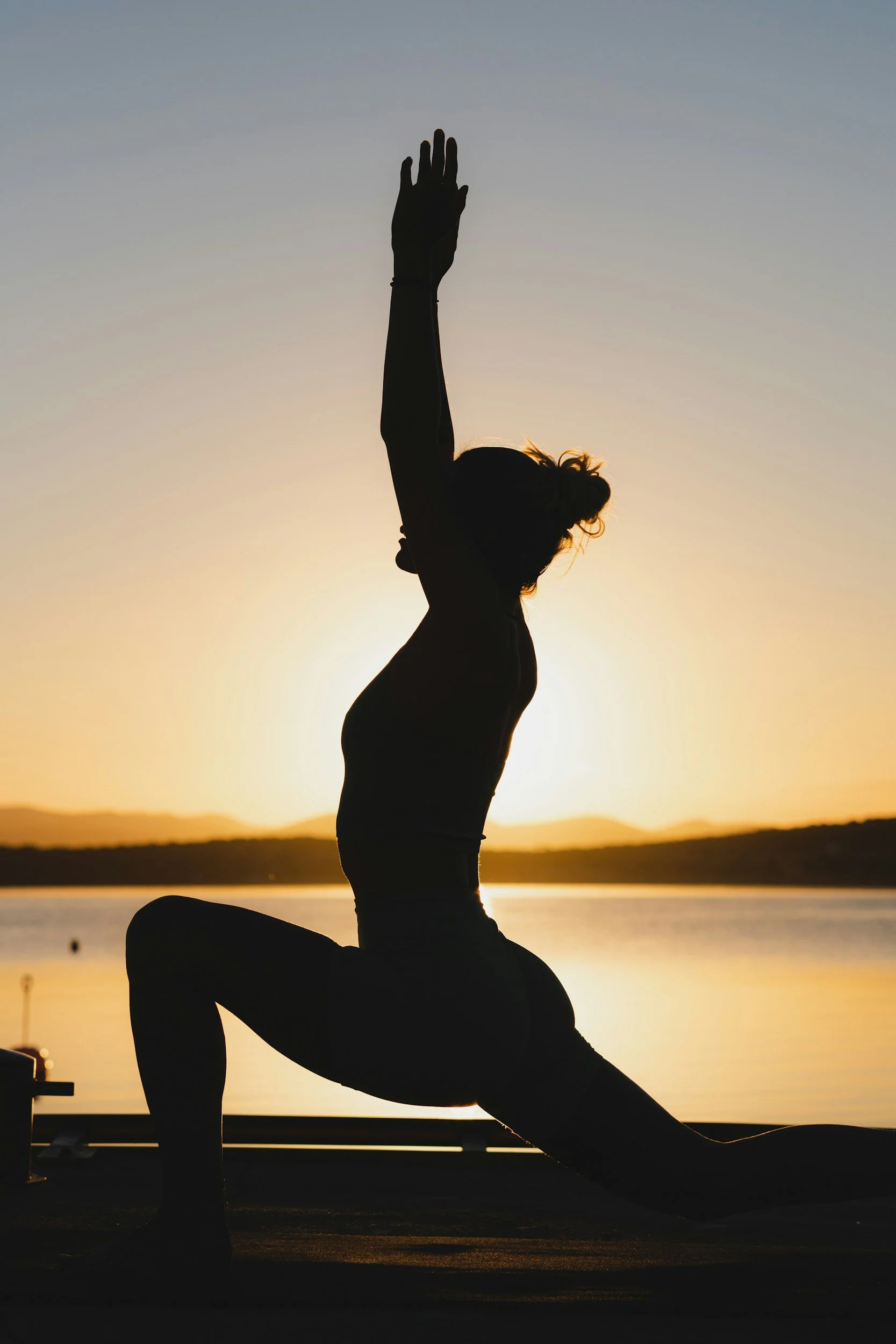 Silhouette of a woman practicing yoga outdoors at sunset, with a body of water and distant hills in the background.