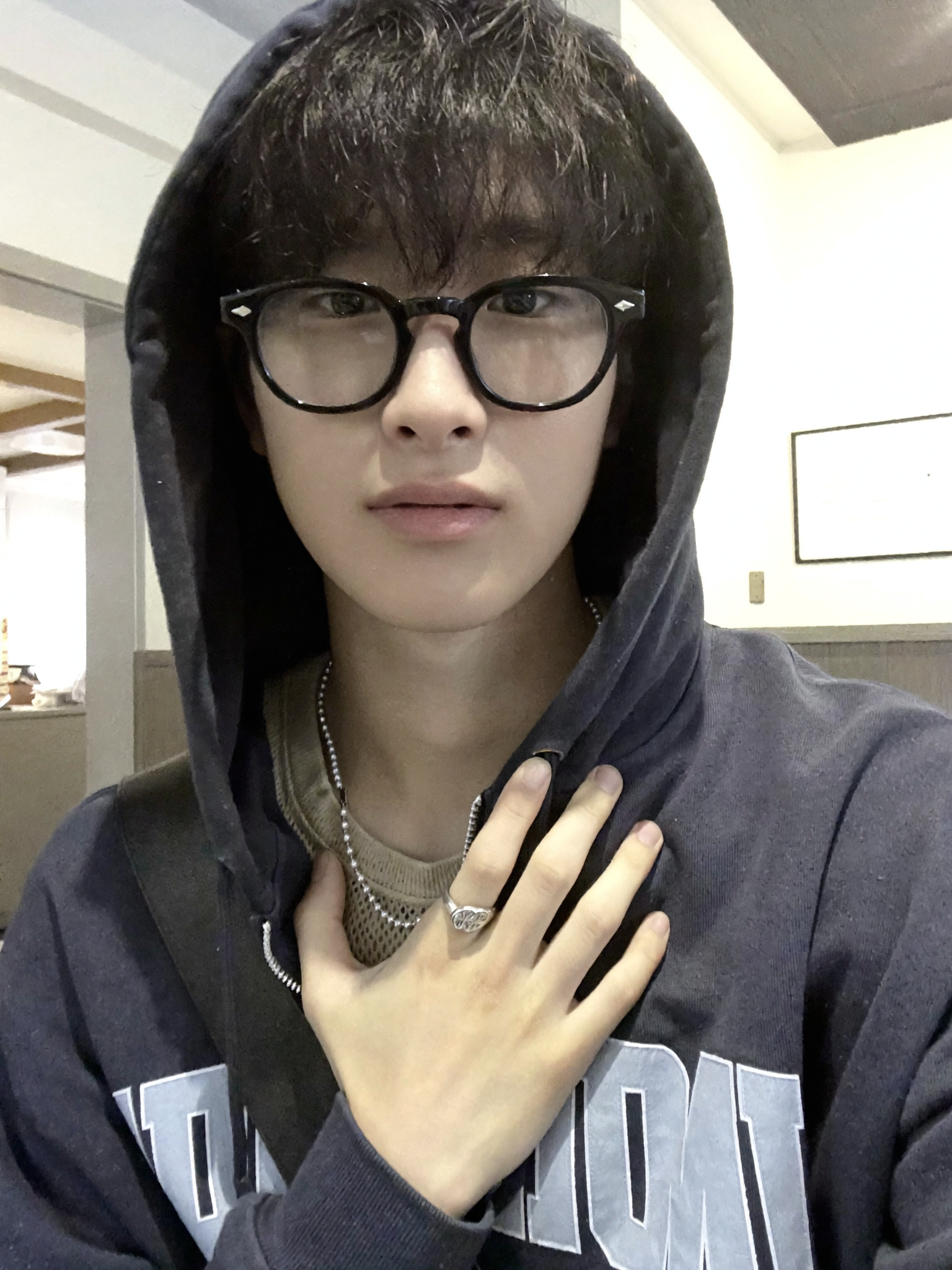 A young person with short dark hair, wearing glasses and a grey hoodie with the hood up, holding their hand over their chest. Background appears to be a casual indoor setting.