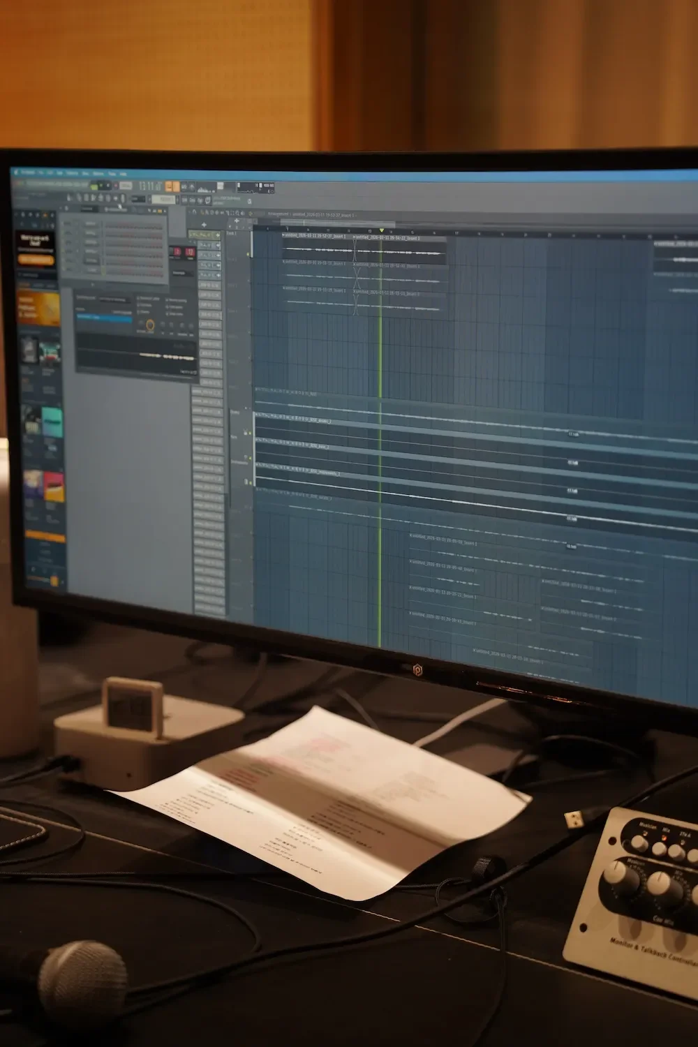 Computer monitor displaying music production software with multiple audio tracks, a microphone on a stand, and a mixing device on the table.