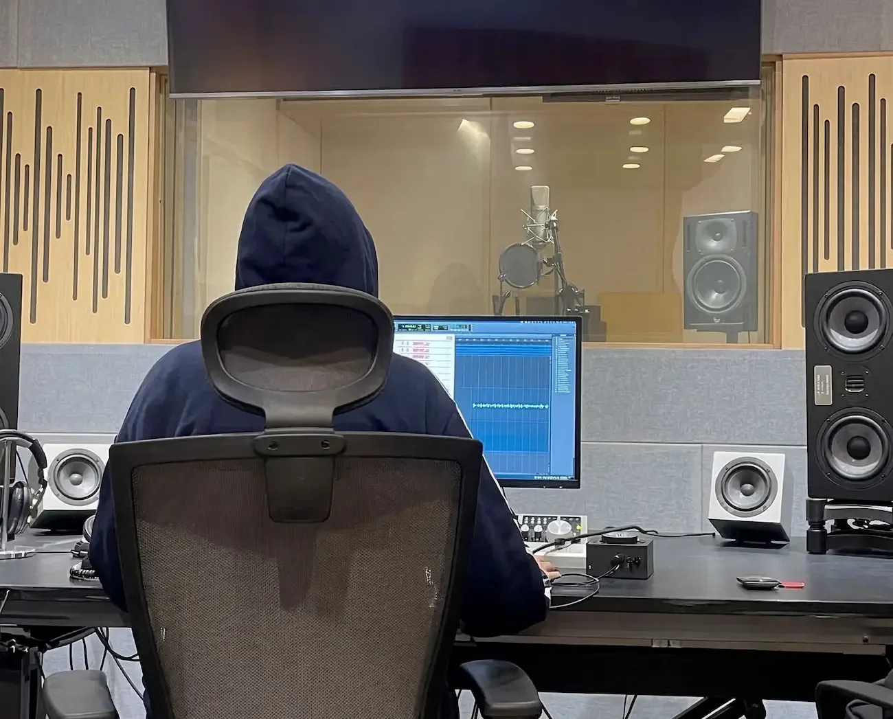 Person wearing a hoodie sitting in a recording studio in front of a computer monitor with audio editing software, facing a glass window into a separate recording booth with a microphone.