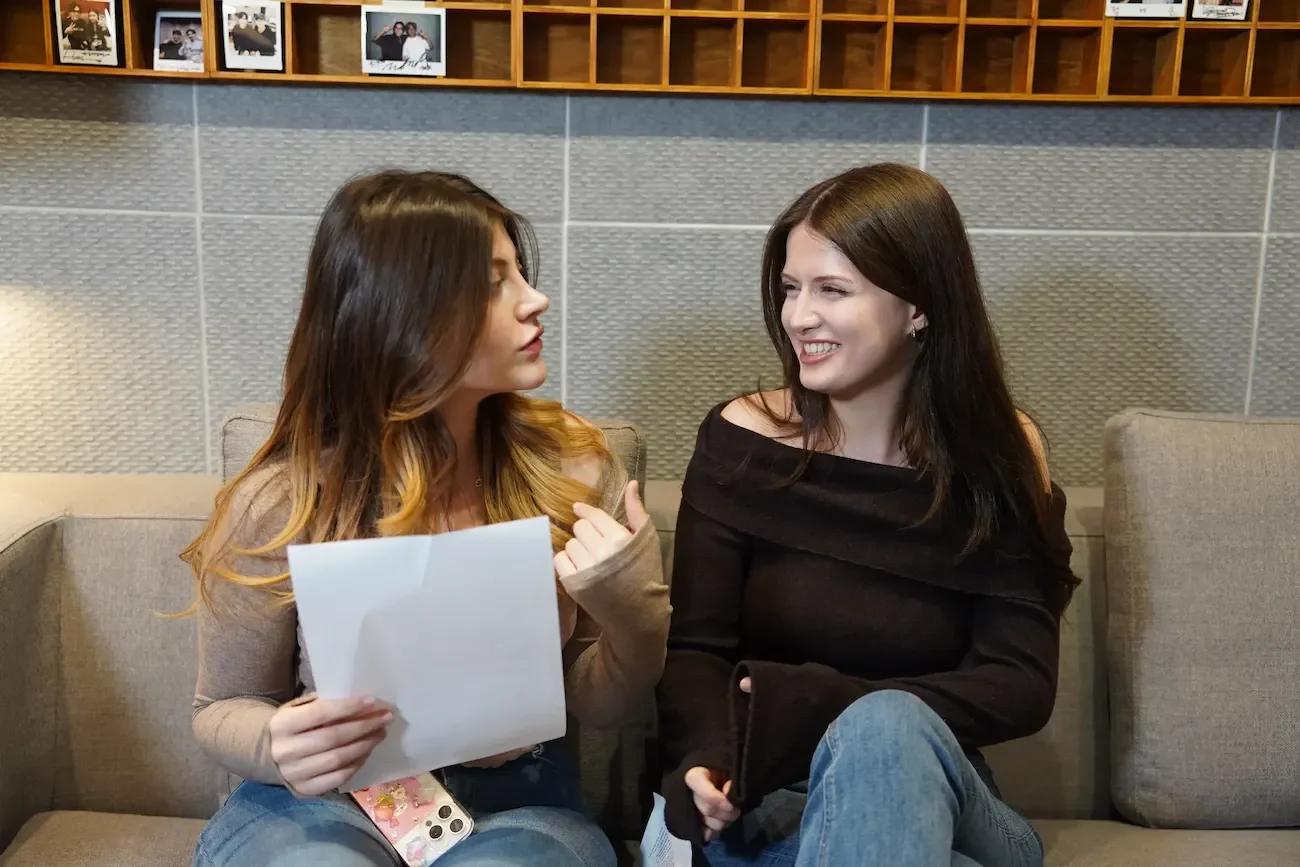 Two women sitting on a beige couch having a conversation, one holding a paper and the other smiling, with framed photos on a wooden shelf above them.