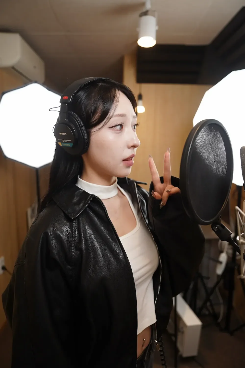 Young woman in a recording studio, wearing headphones and a black leather jacket, posing with a peace sign near a microphone in front of a pop filter.