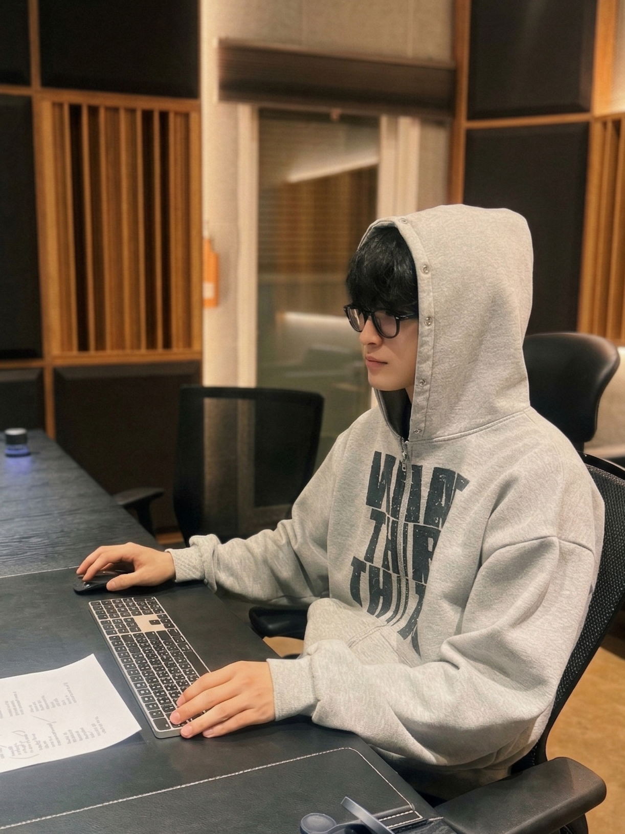Young man with black hair and glasses wearing a grey hoodie, sitting at a desk using a computer in a recording studio.