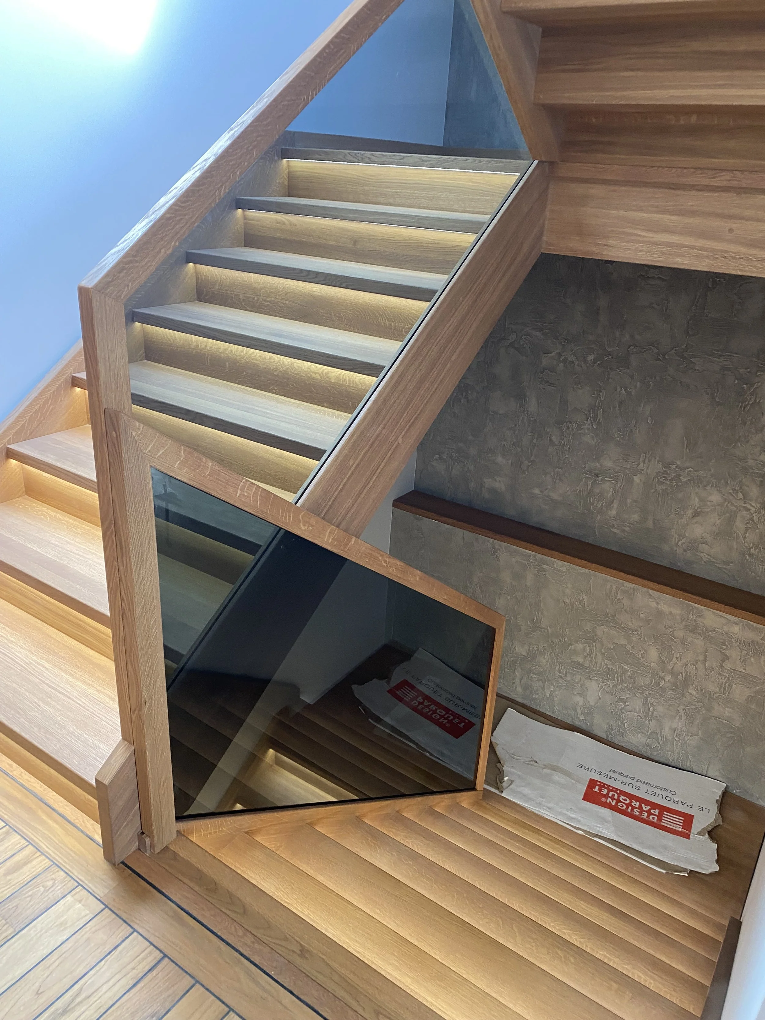 Mediniai laiptai su stiklo plokštėmis ir integruotu apšvietimu, vedantys žemyn į apatinį aukštą.