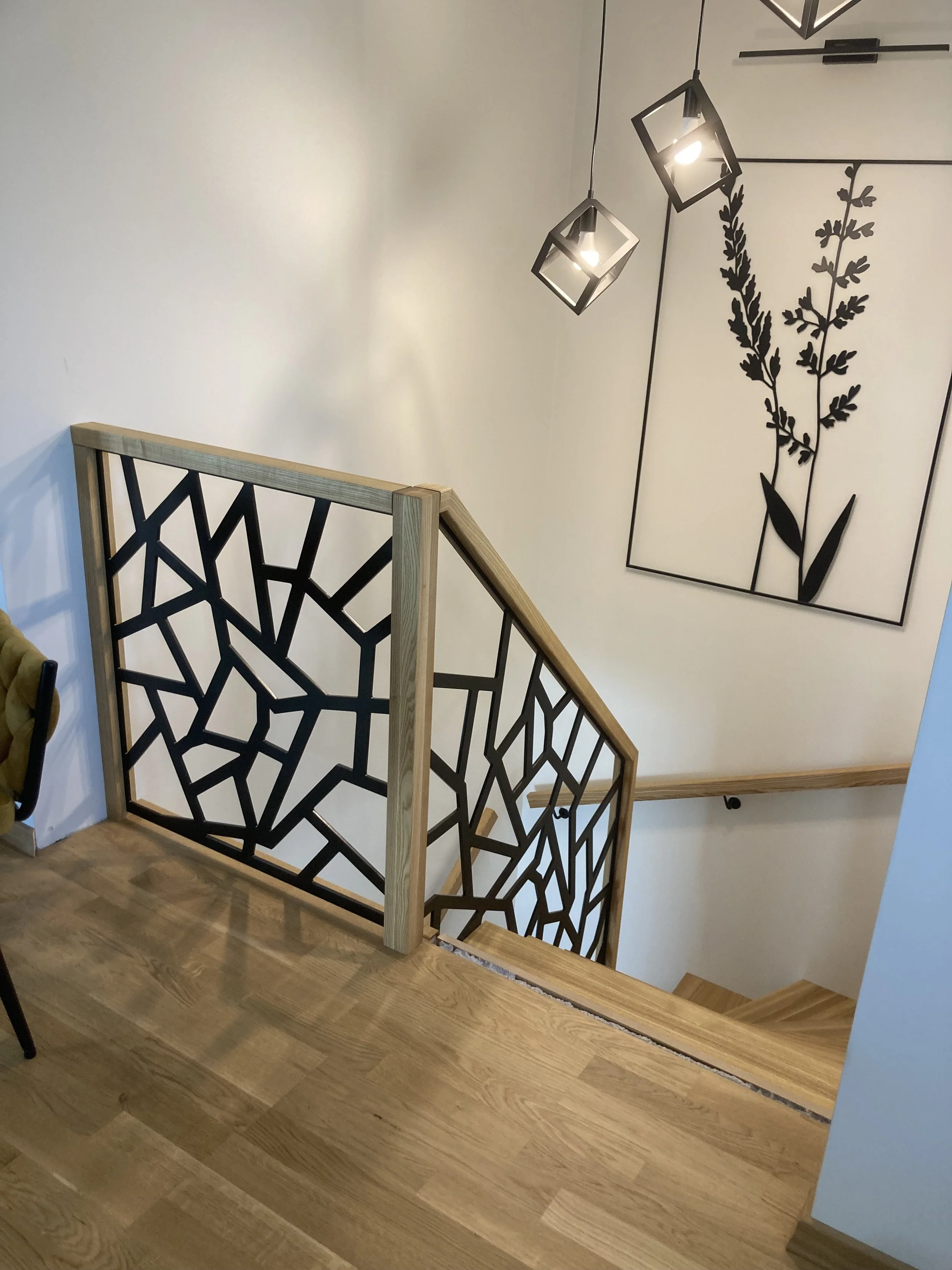 Mediniai laiptai su geometriniais juodo metalo turėklais, kabančiais geometriniais šviestuvais, botaniniu siluetu dekoruota siena ir šviesiomis medinėmis grindimis.