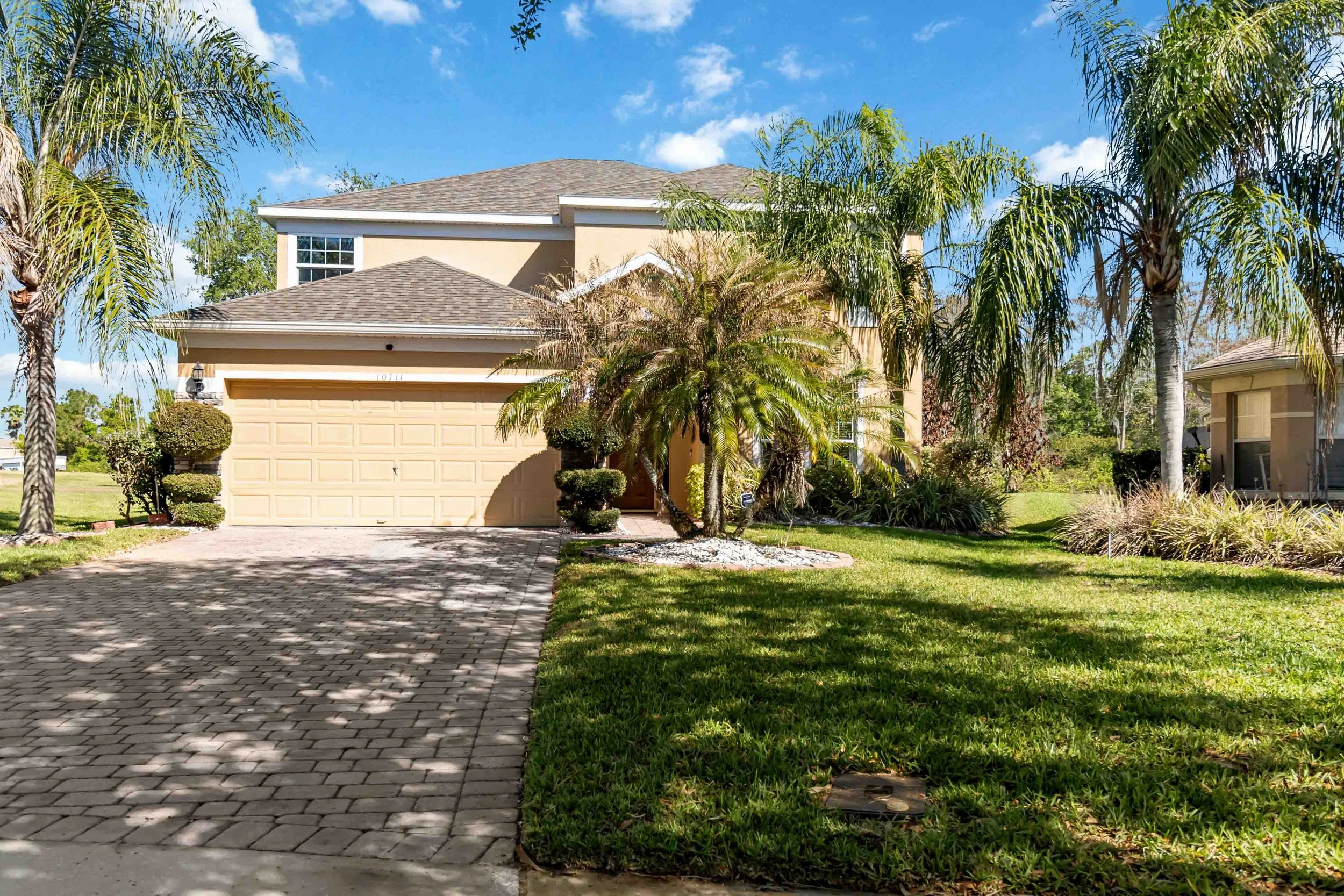 A beige two-story house with a garage, surrounded by tall palm trees, manicured shrubs, and a green lawn, under a blue sky with some clouds.