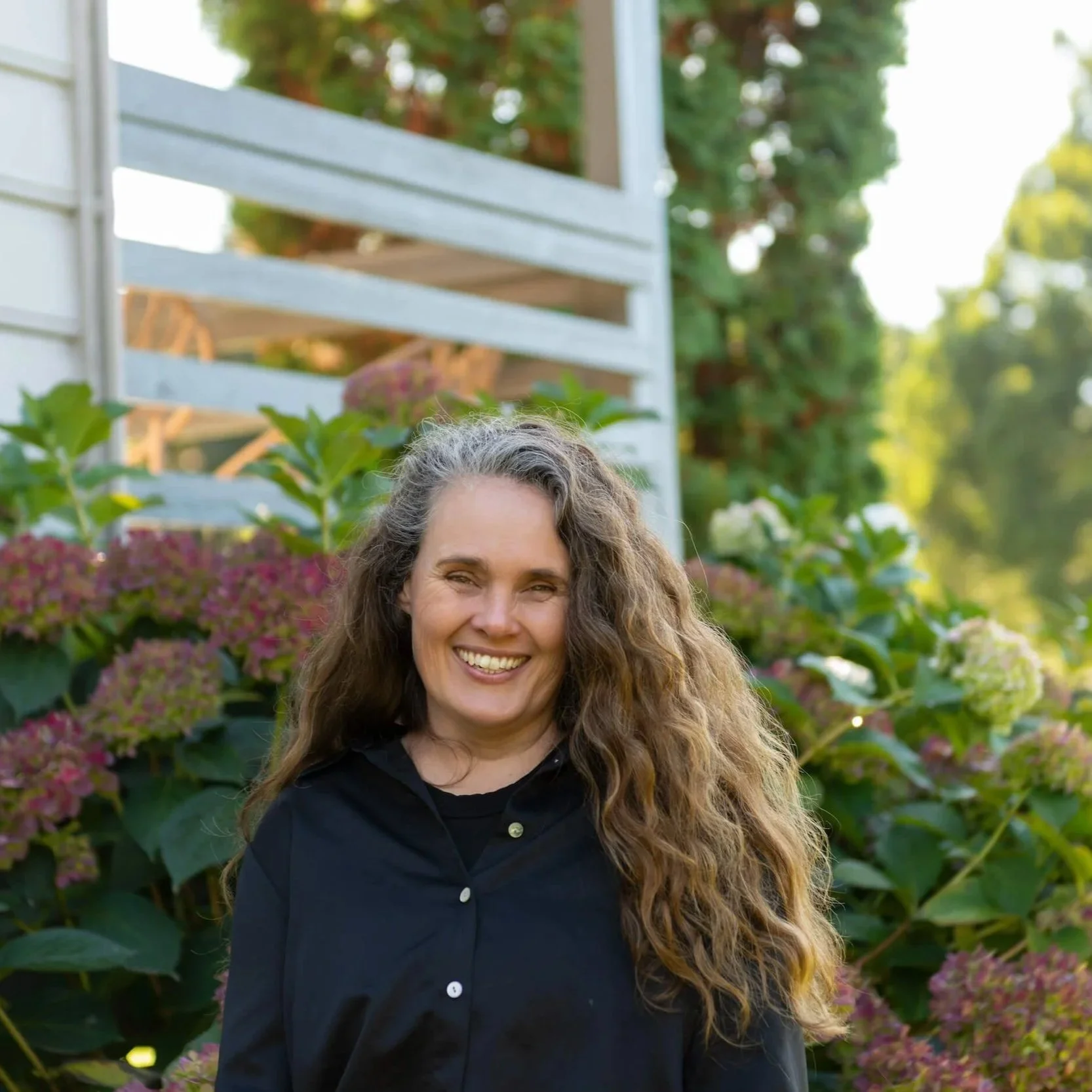 A woman with long, curly, gray hair, smiling, standing outdoors in front of pink flowers and green foliage, with a white wooden or metal structure in the background.