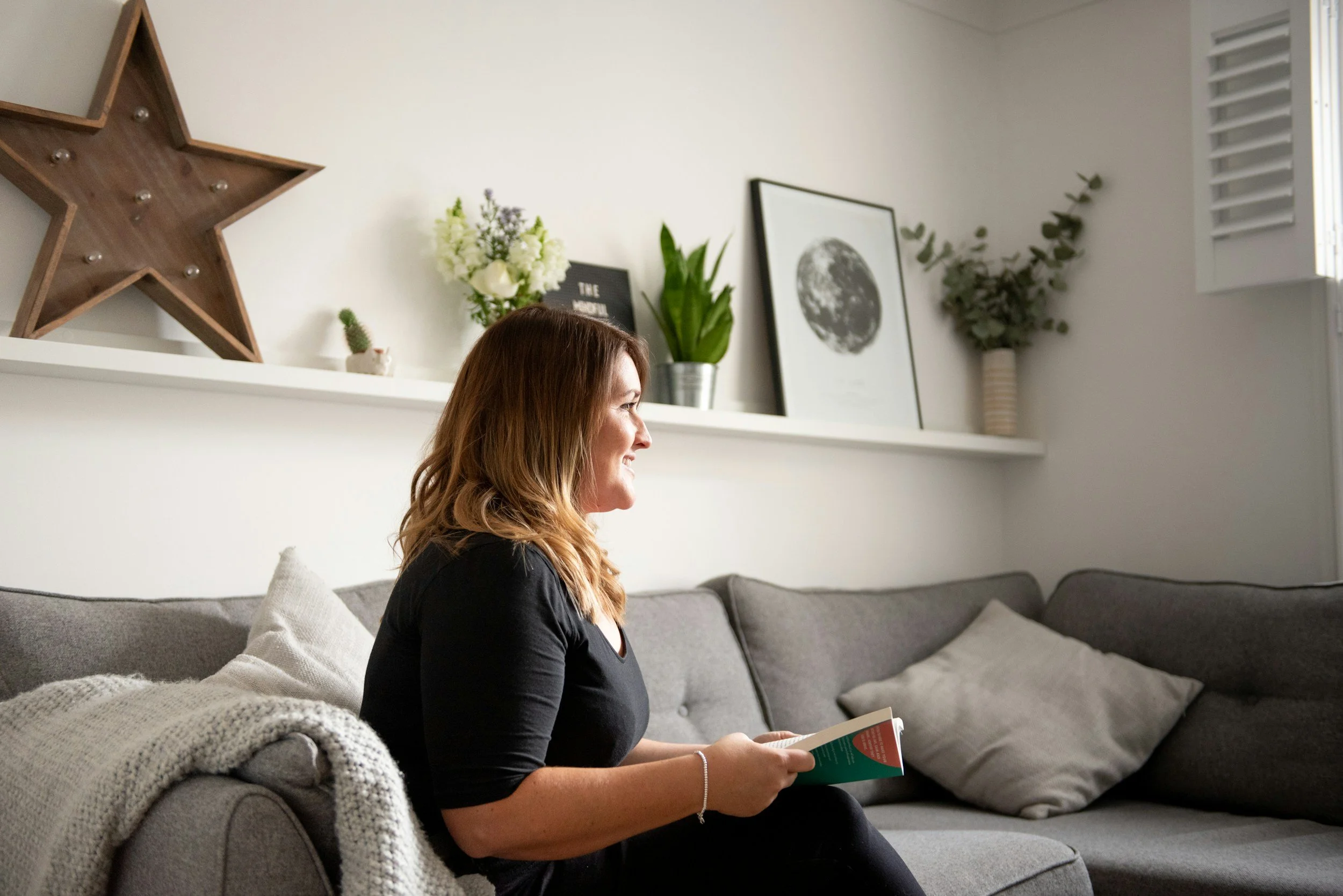 A woman with brown hair, wearing a black shirt, sitting on a gray sofa with a book in her hands. The background features a white wall with decorative items including a large wooden star, framed black-and-white moon print, plants, and a small cactus.