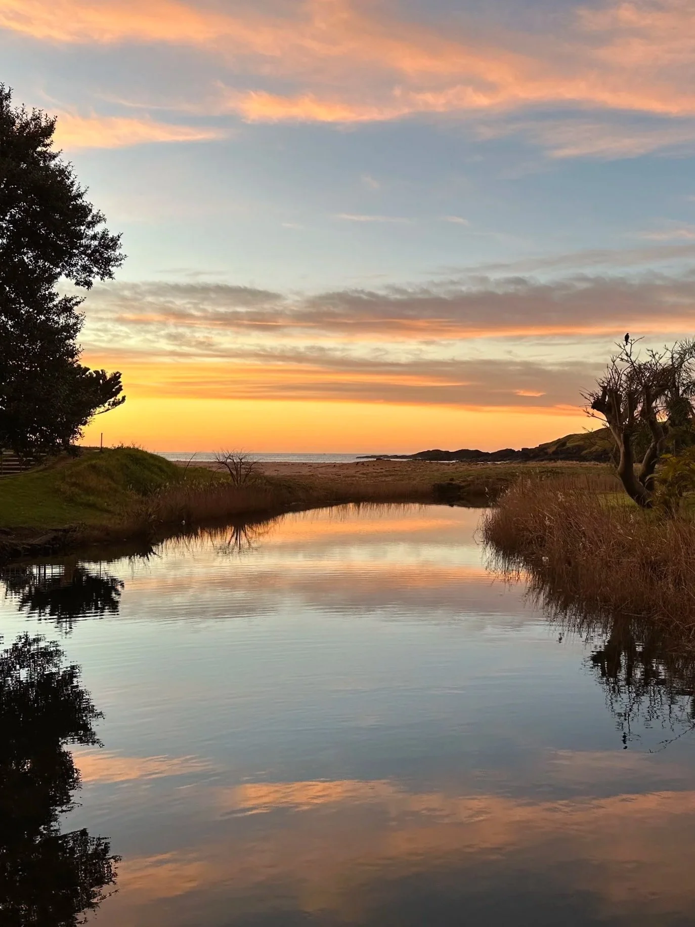 Sunset over a calm body of water, with colorful clouds in the sky, trees on either side, and the water reflecting the sky's colors.