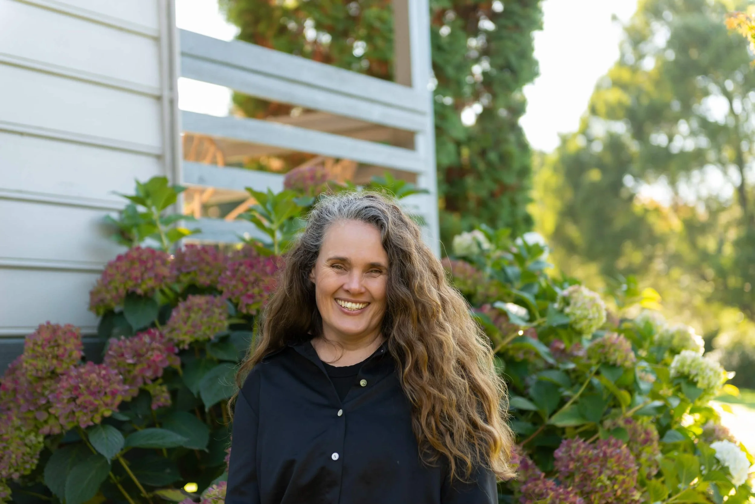 Karina, Neerim South naturopath outdoors in front of blooming hydrangea bushes with a house and trees in the background.