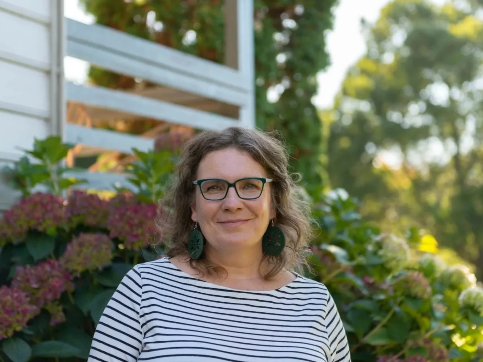 Ness, Neerim South counsellor wearing glasses and earrings smiling outdoors with green plants and pink flowers in the background.