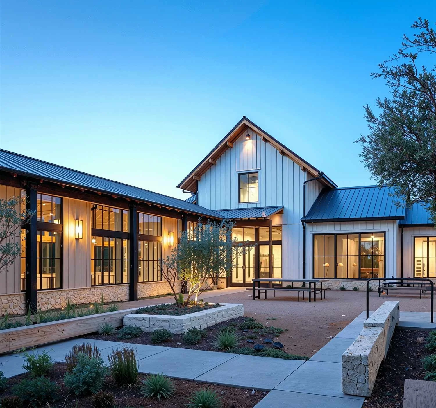 Modern style building with large windows and metal roof, surrounded by landscaped yard and patio, at dusk.
