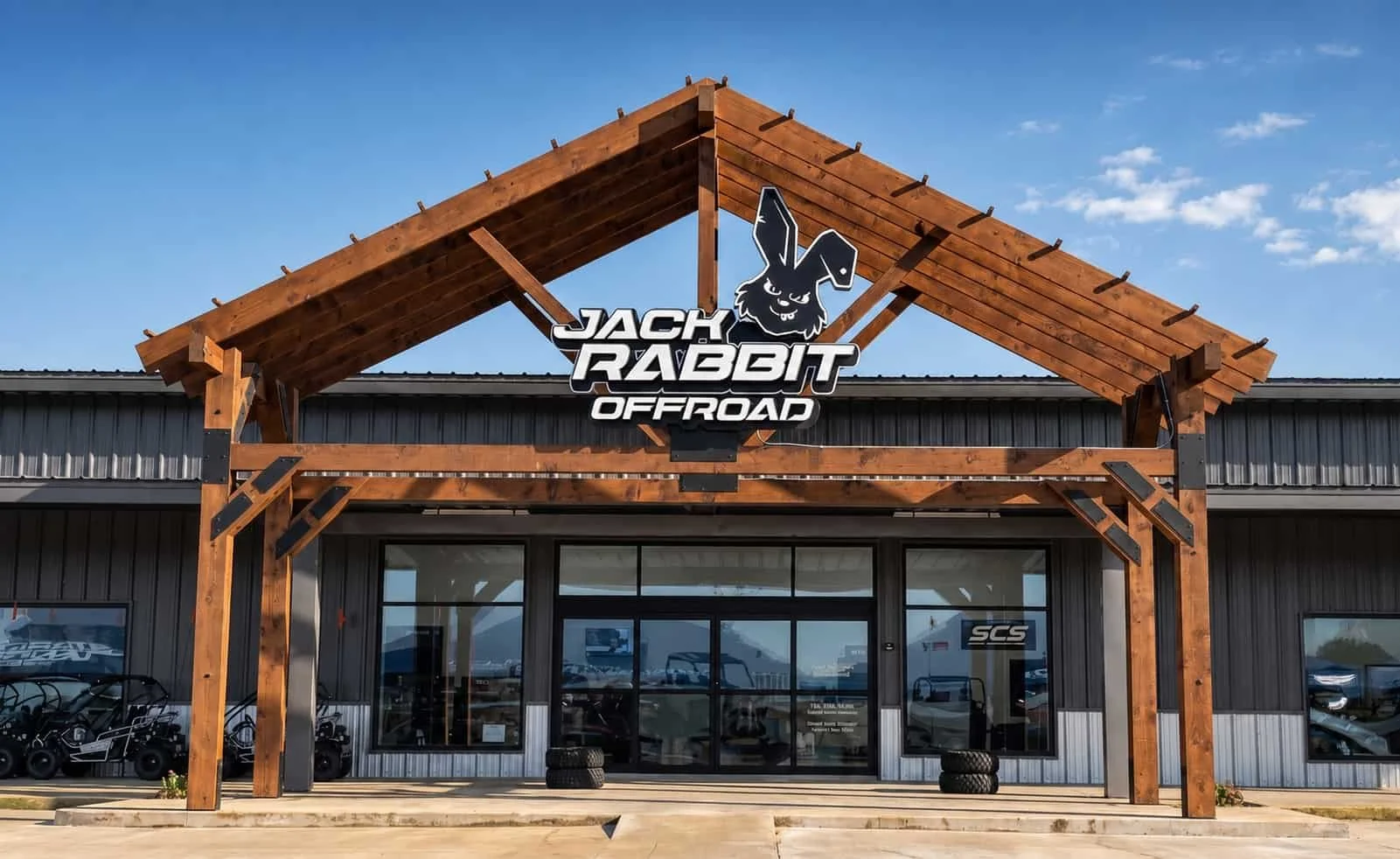 Front view of a building with a black metal exterior and large glass windows. A wooden structure with a gabled roof frames the entrance, featuring a sign with a rabbit logo and the text 'Jack Rabbit Offroad' above the entrance.