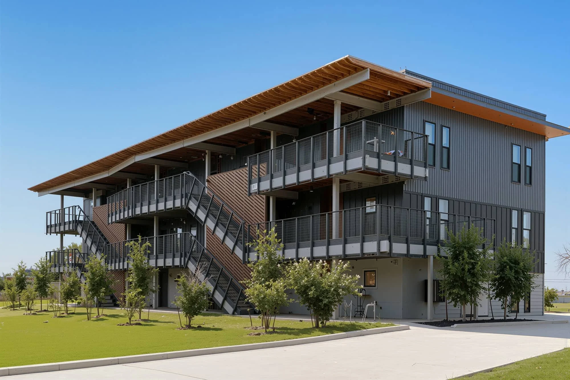 Modern multi-story building with metal siding, balconies, and external stairs, surrounded by green lawn and young trees under a clear blue sky.