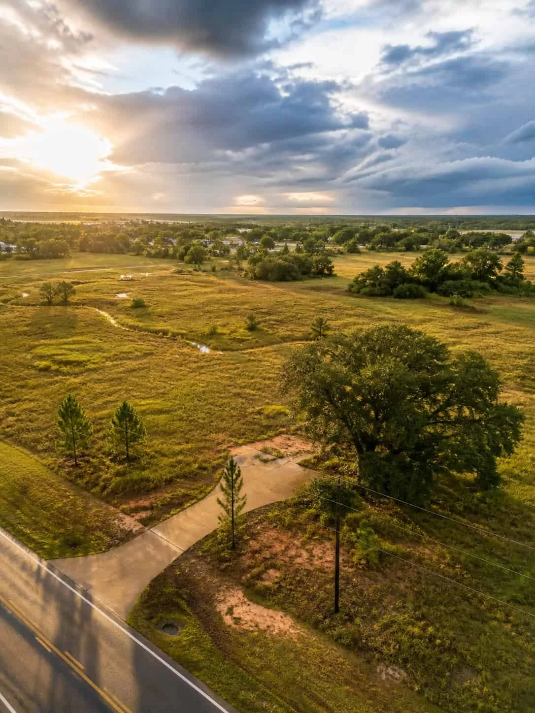 Aerial view of a rural landscape during sunset, showing open fields, scattered trees, a walkway curving through the area, a road at the bottom, and a stormy sky with dark clouds and sunlight breaking through.