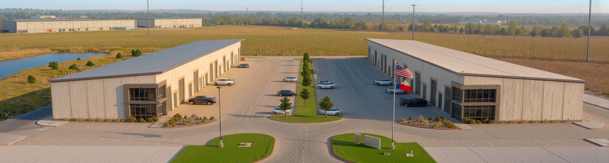 Aerial view of a modern commercial building with parking lots, trees, and American and Texas flags in front, surrounded by open land and a small pond.