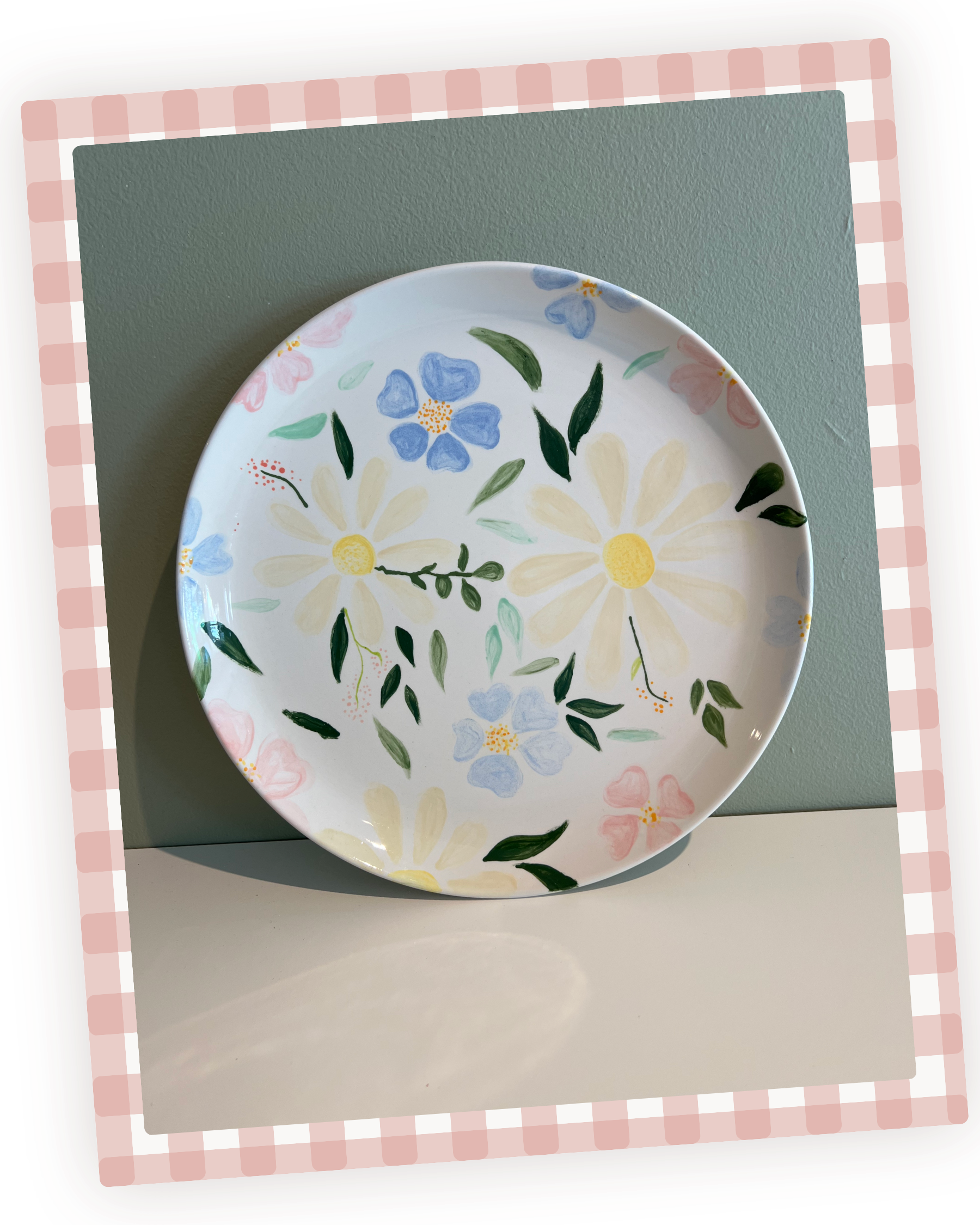 En rund porcelæns tallerken med blomsterdesign i blå, gul, grøn og pink, placeret på en grøn underkop og en hvid bordplade med en pink-hvid tjekket borddækning rundt om.