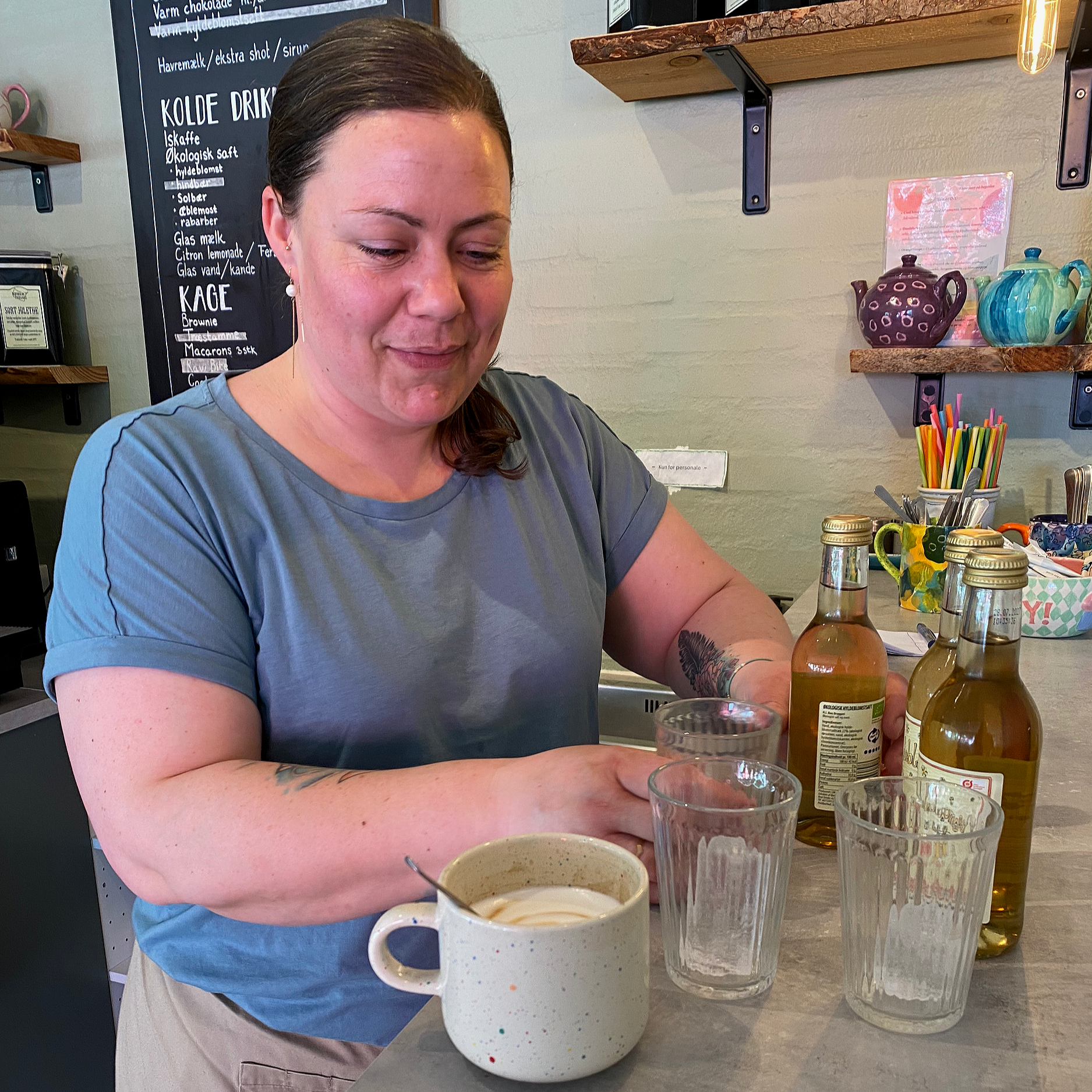 En kvinde smiler i en cafe, omgivet af forskellige drikke, herunder saftevand og kaffe, med et sort tavle med menu i baggrunden.