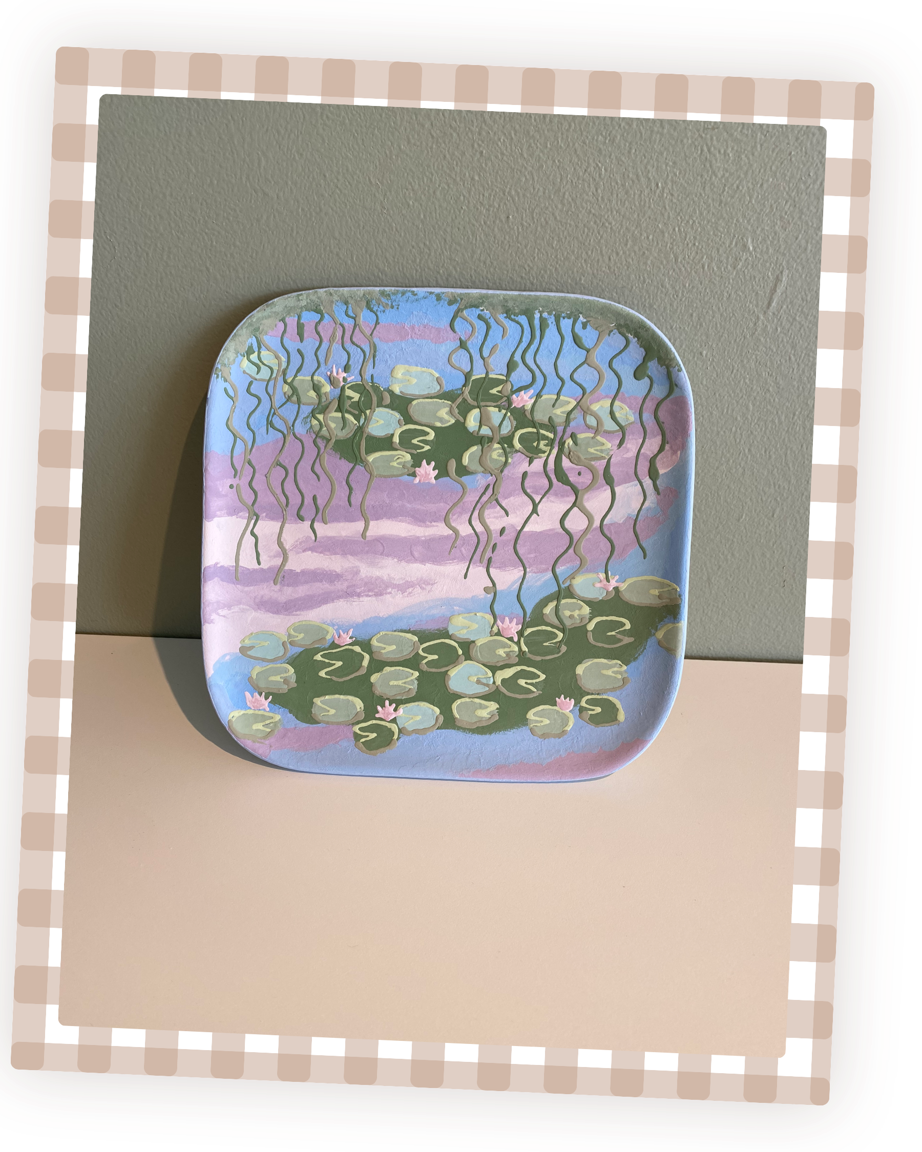 Billedet viser en painted tallerken med et miniature vandfald af vandplanter, papirfugle og blomster, placeret på en beige overflade.