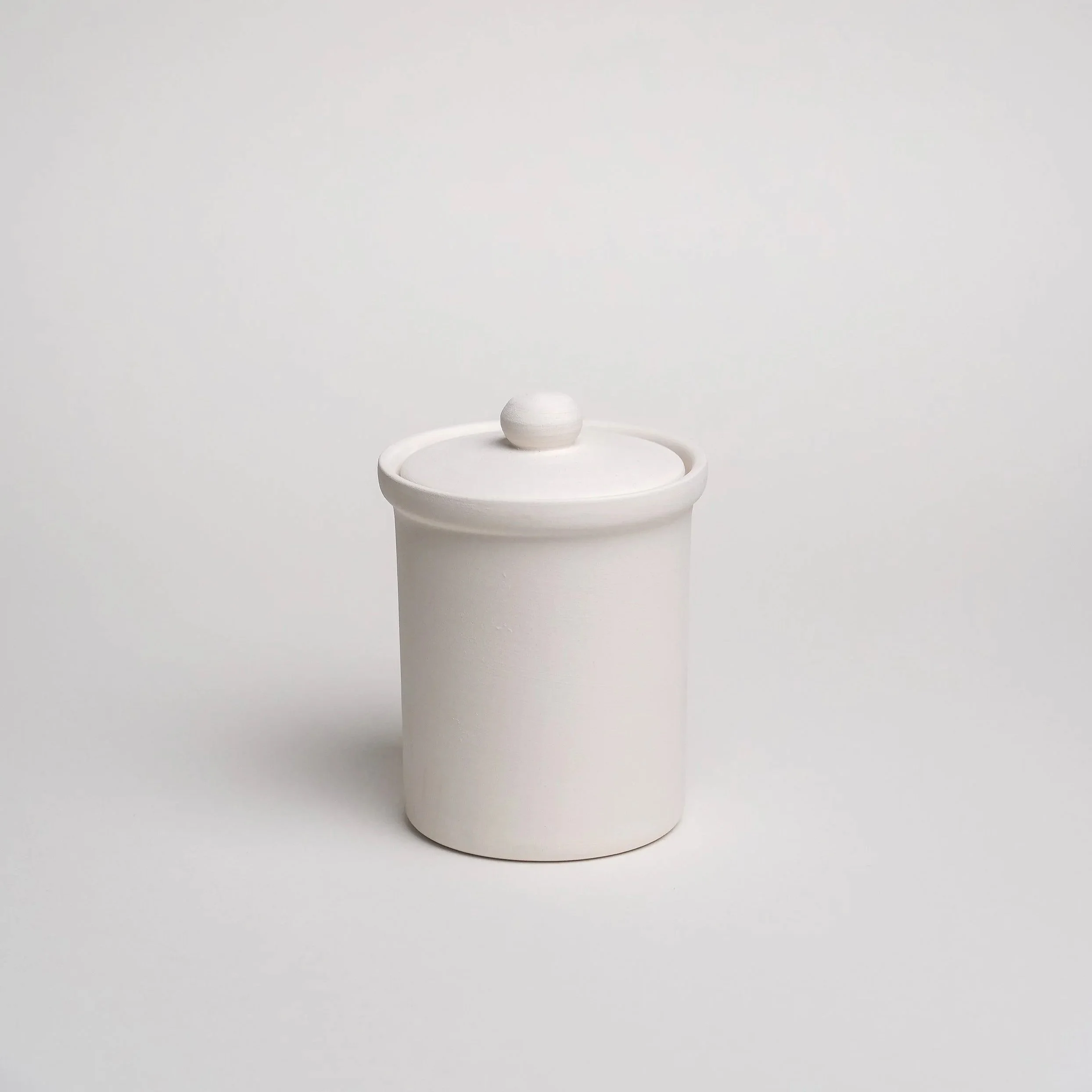 En hvid keramisk vase med en kugleformet top, placeret på en høj, hvid, cylindrisk sokkel mod en neutral baggrund.