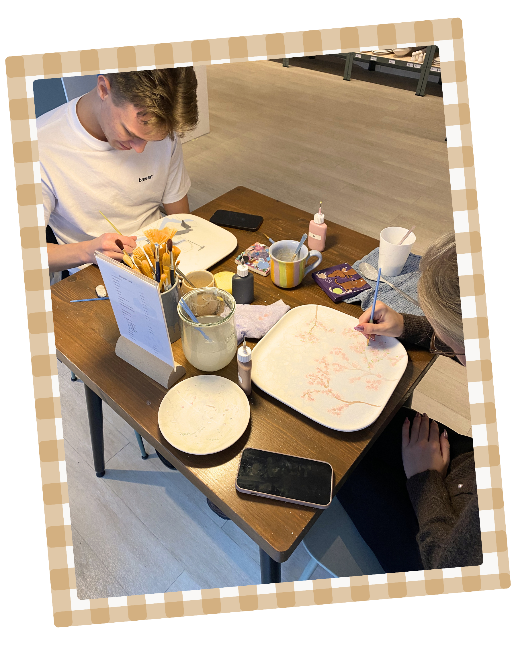 En ung mand sidder ved et bord, der er dækket med malingudstyr, vaser med maling, tallerkener med malerier og en kop med en ske. Han male på en stor papirplade. En telefon og en menu med drikkevarepriser står på bordet.