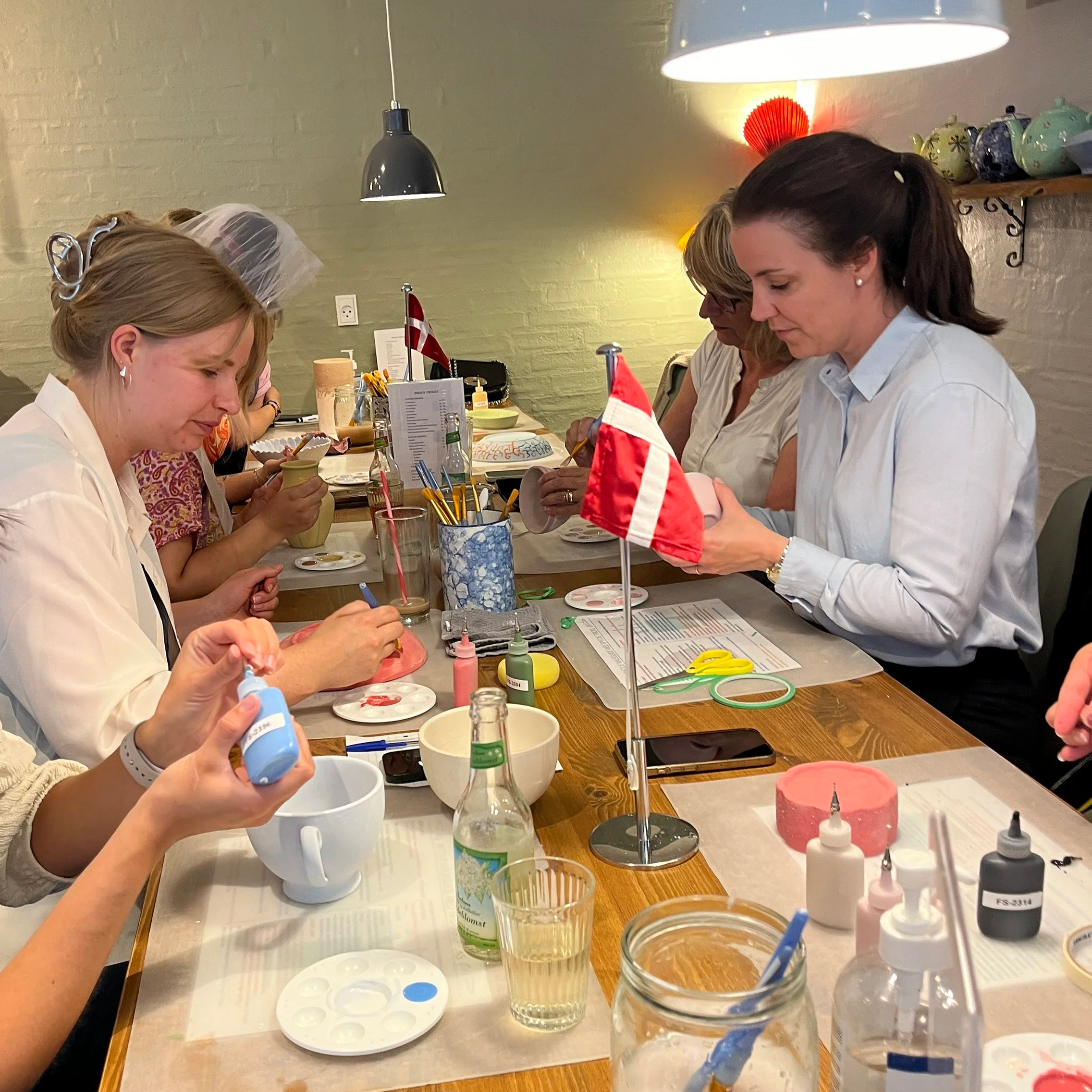 Gruppe kvinder, der sidder ved et bord og maler med porcelænsfarver, omringet af malerpaletter, pensler og porcelænsgenstande