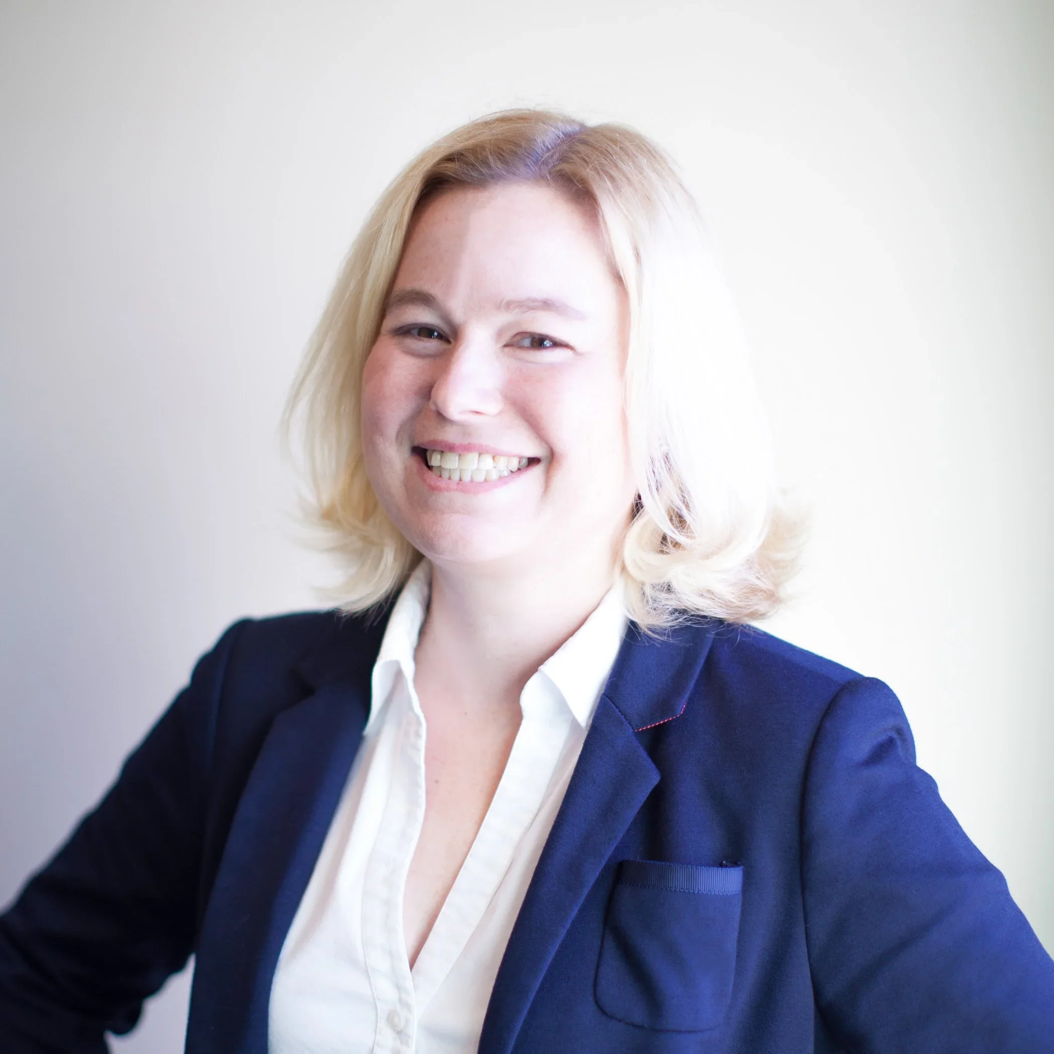 A woman with blonde hair smiling, wearing a white shirt and dark blazer, against a plain light background.