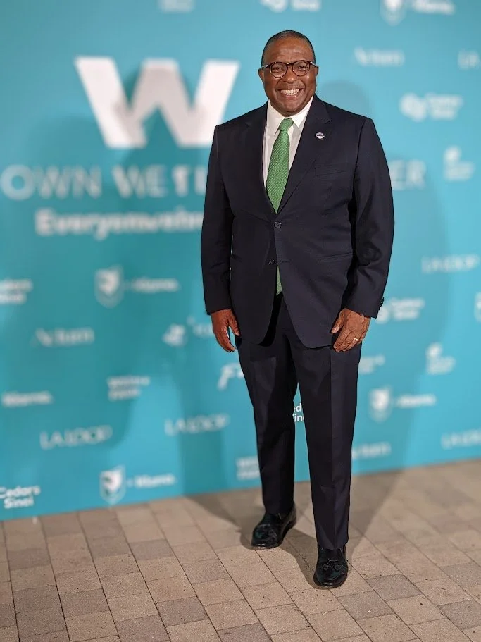 A man in a dark suit with a white shirt and green tie standing and smiling in front of a turquoise backdrop with white logos and text.