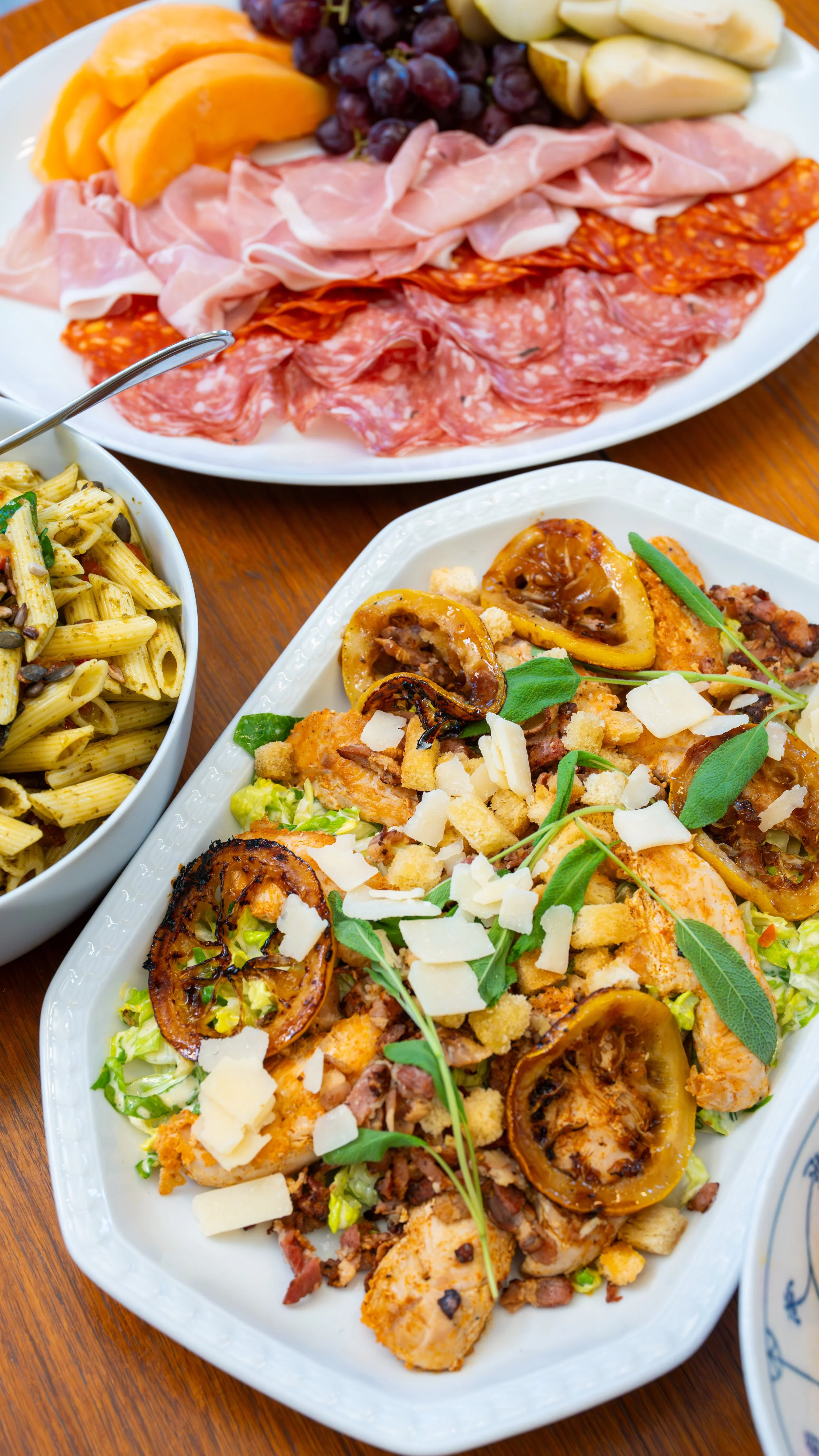 A platter of assorted sliced fruits, including cantaloupe, grapes, and apple slices, with a variety of cured meats below. A separate dish contains pasta salad with olives and herbs, and another dish features roasted vegetables and grilled chicken topped with cheese and basil.