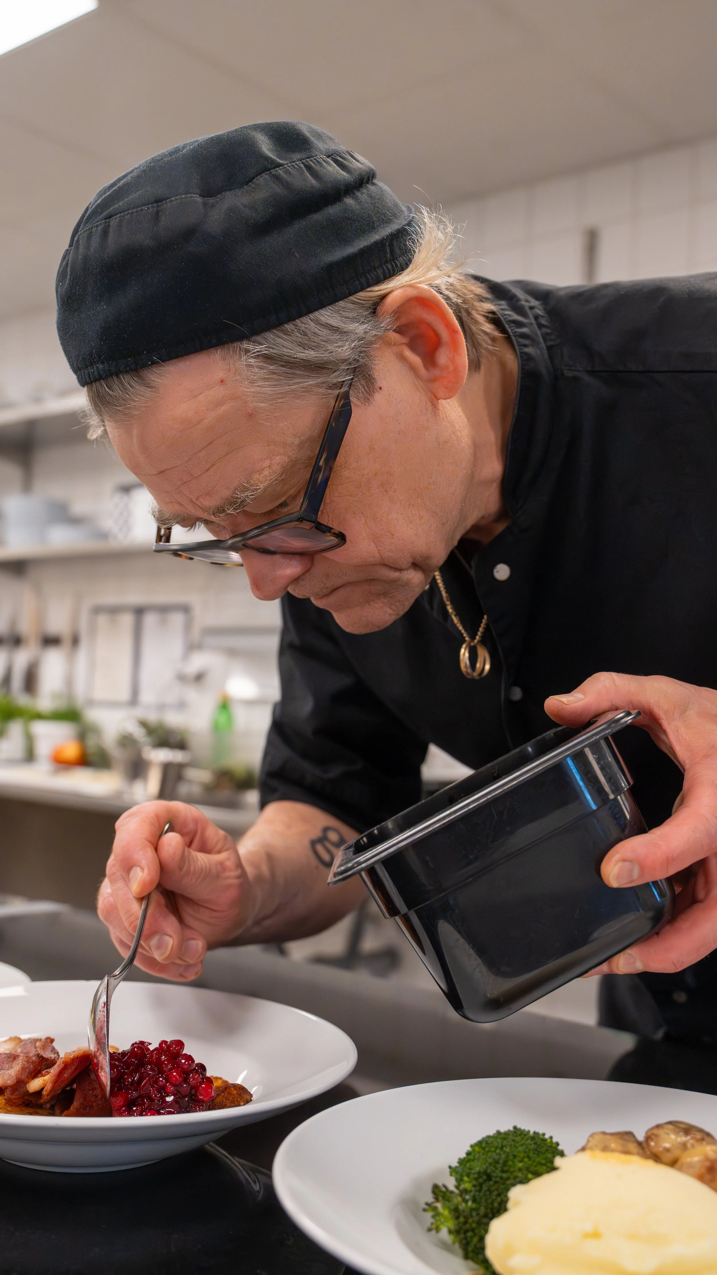 A chef in a black uniform and hat garnishing dishes in a professional kitchen. The chef is finishing plates of food, which include a dish with cranberries and another with broccoli and mashed potatoes.
