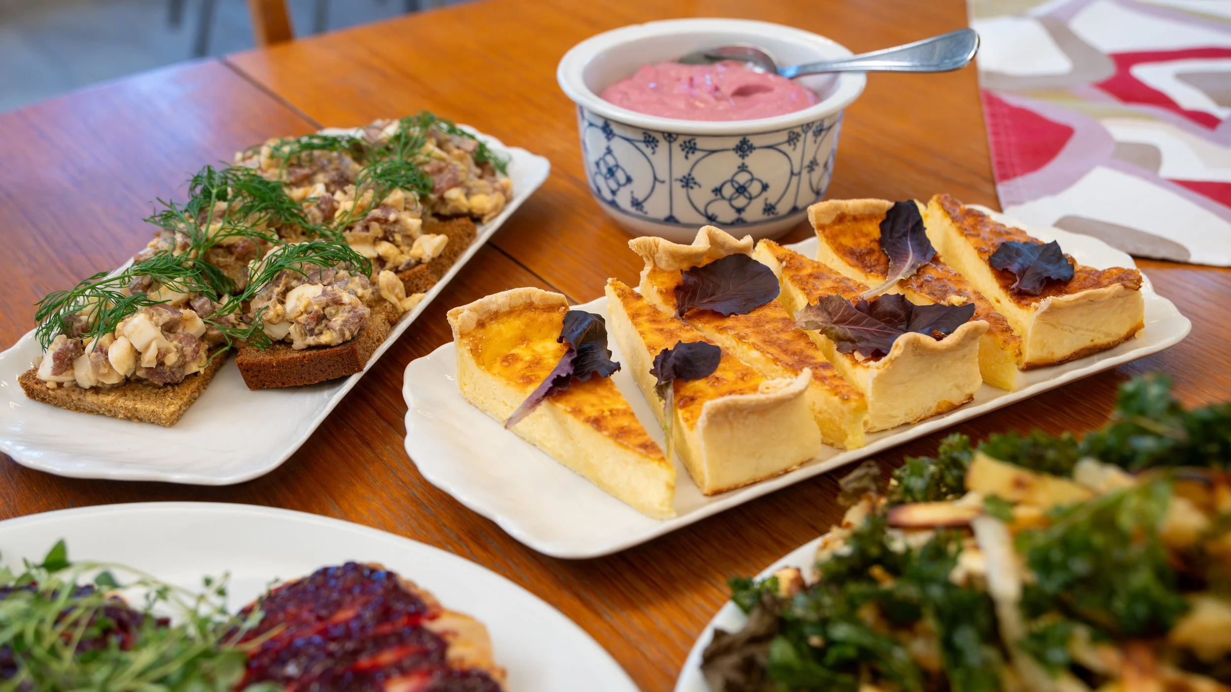 Assorted dishes on a wooden table, including a slice of bread with a creamy topping garnished with herbs, slices of quiche with dark leaves on top, and a bowl of pink pudding with a spoon.