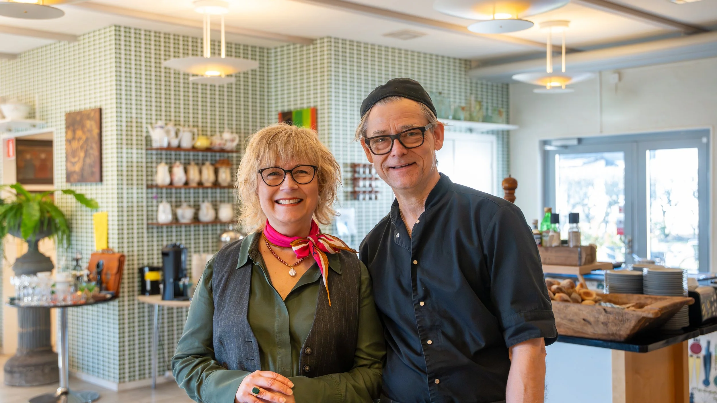 Monika Georgsson and Stefan Georgsson standing together in a home or cafe setting with green checkered wall, shelves with jars, and a counter with baked goods.