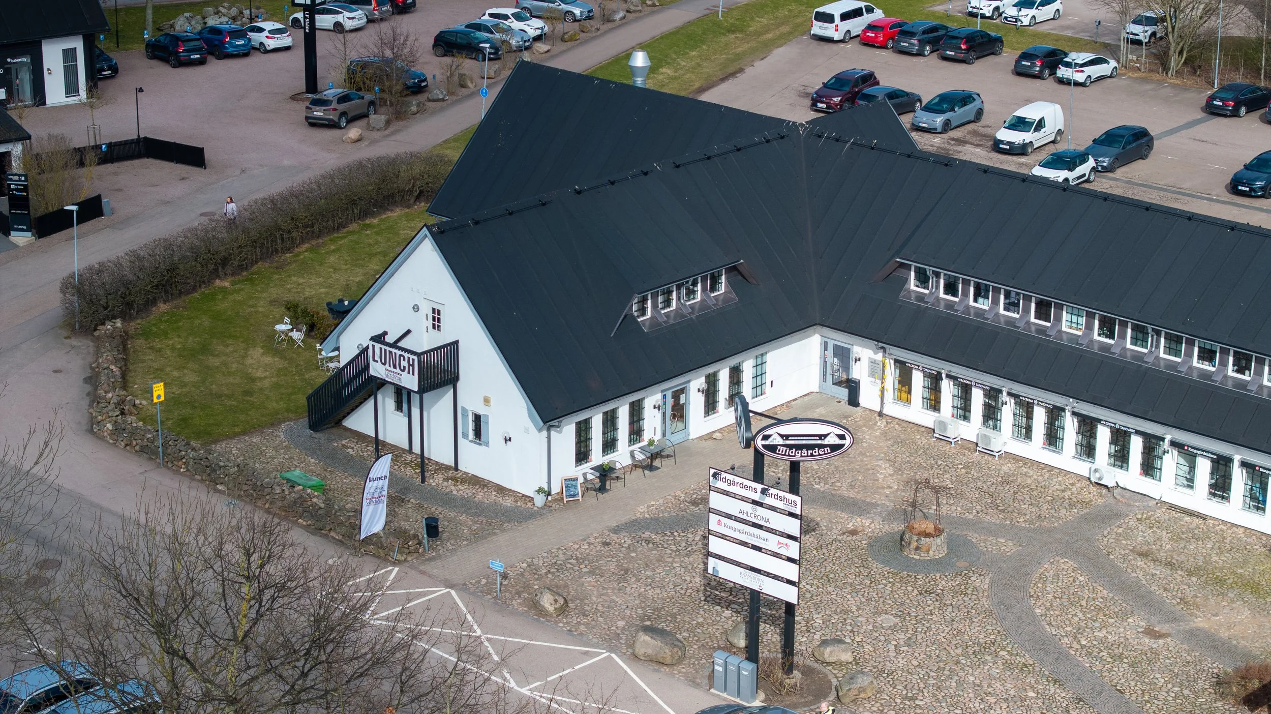 An aerial view of a building with a black roof and white walls, surrounded by parking lots and roads, with a small grassy area and trees nearby.