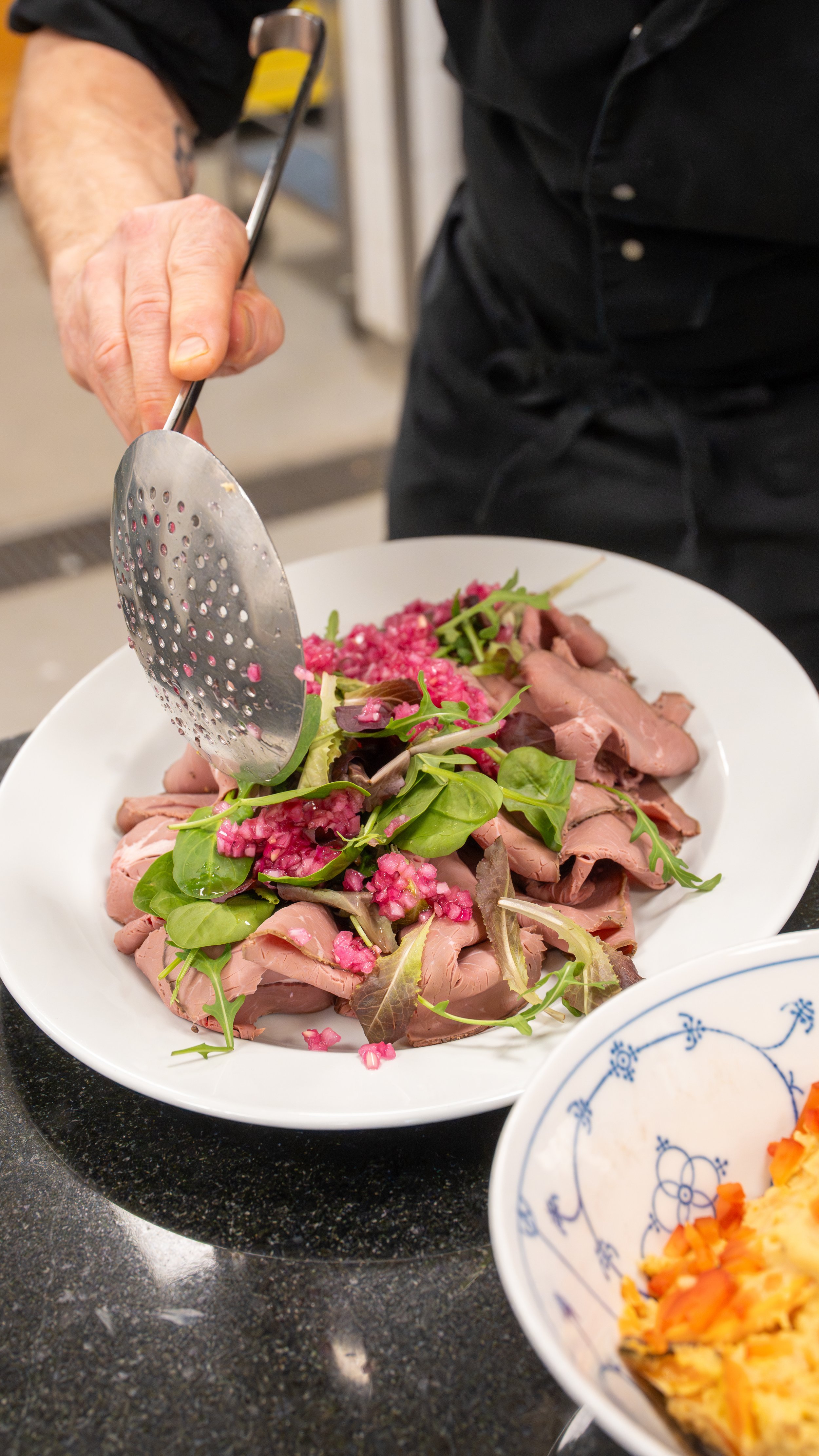 A chef prepares a dish with sliced roast beef, fresh greens, and diced pink onions on a white plate, using a slotted spoon. A blue and white bowl with chopped vegetables is partially visible in the foreground.