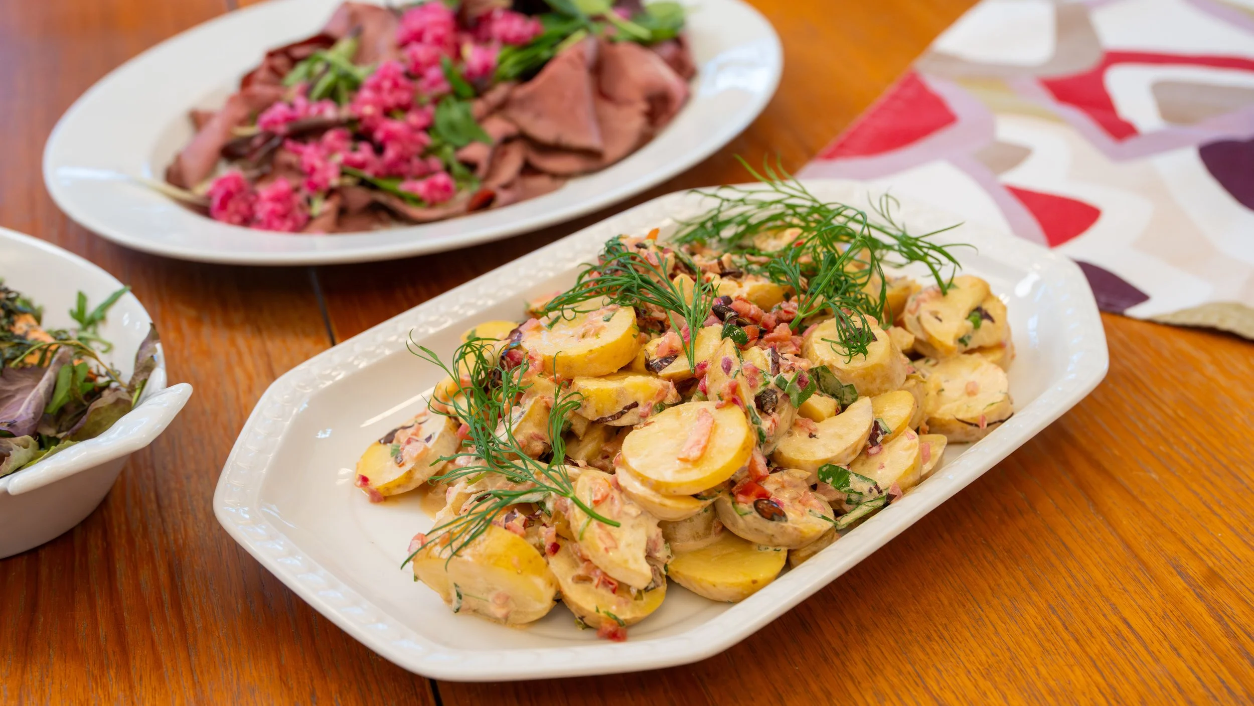 Assorted dishes on white plates including a potato salad garnished with fresh herbs, a meat dish with pink flowers, and a salad with greens, all on a wooden table.