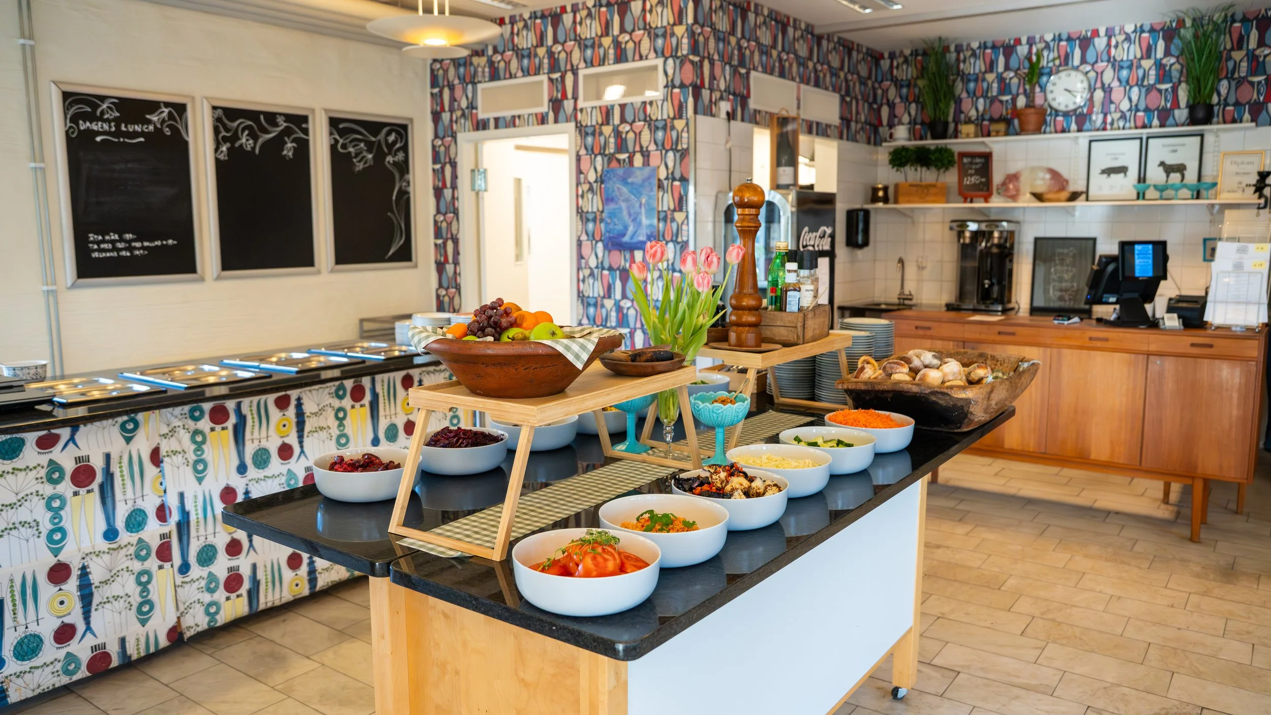 Buffet table with bowls of salad, fruits, and toppings inside a restaurant with colorful wallpaper, chalkboards, and kitchen area in the background.