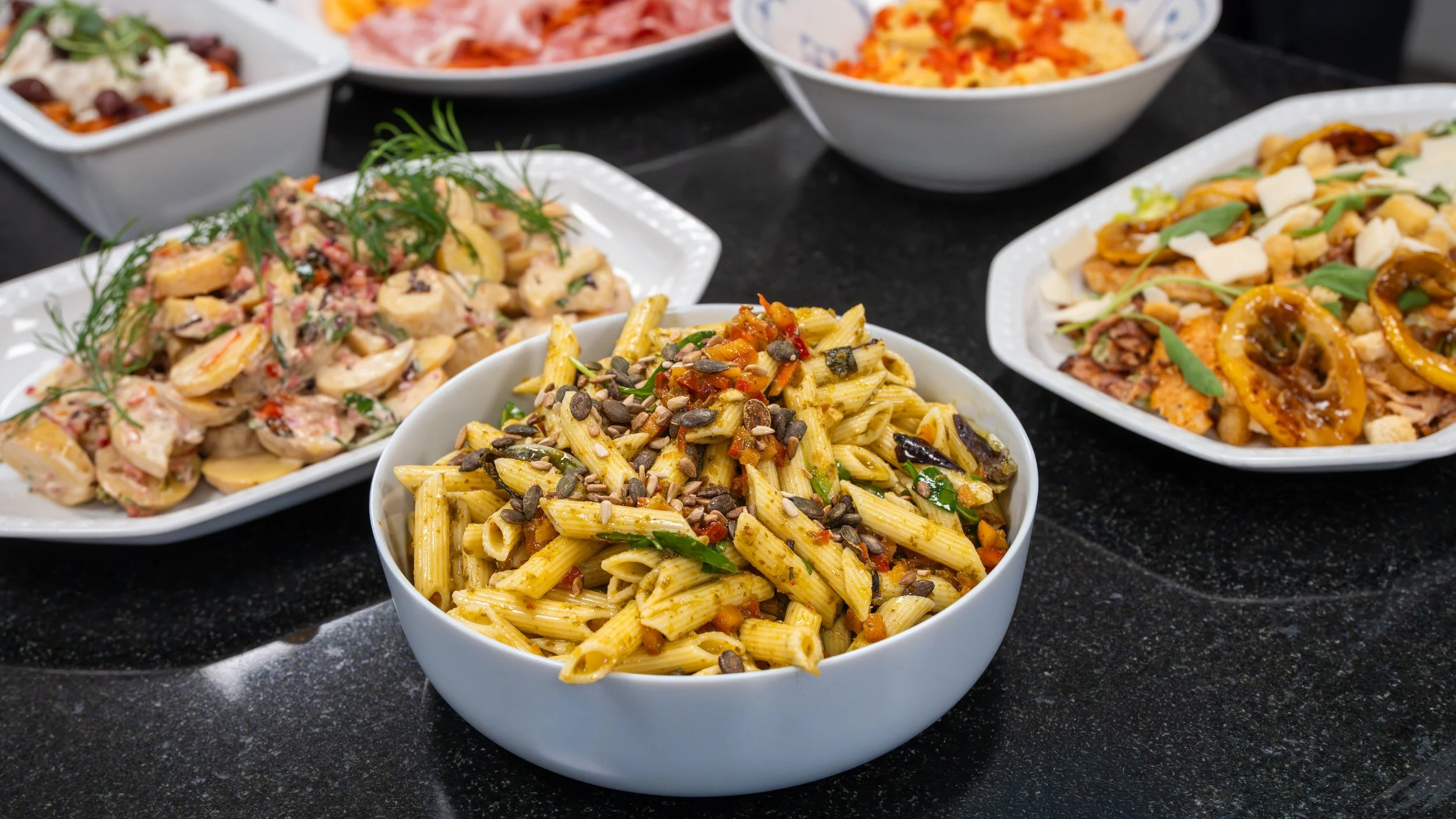 Bowl of pasta with sunflower seeds on top, surrounded by various bowls of salads and dishes on a black table.