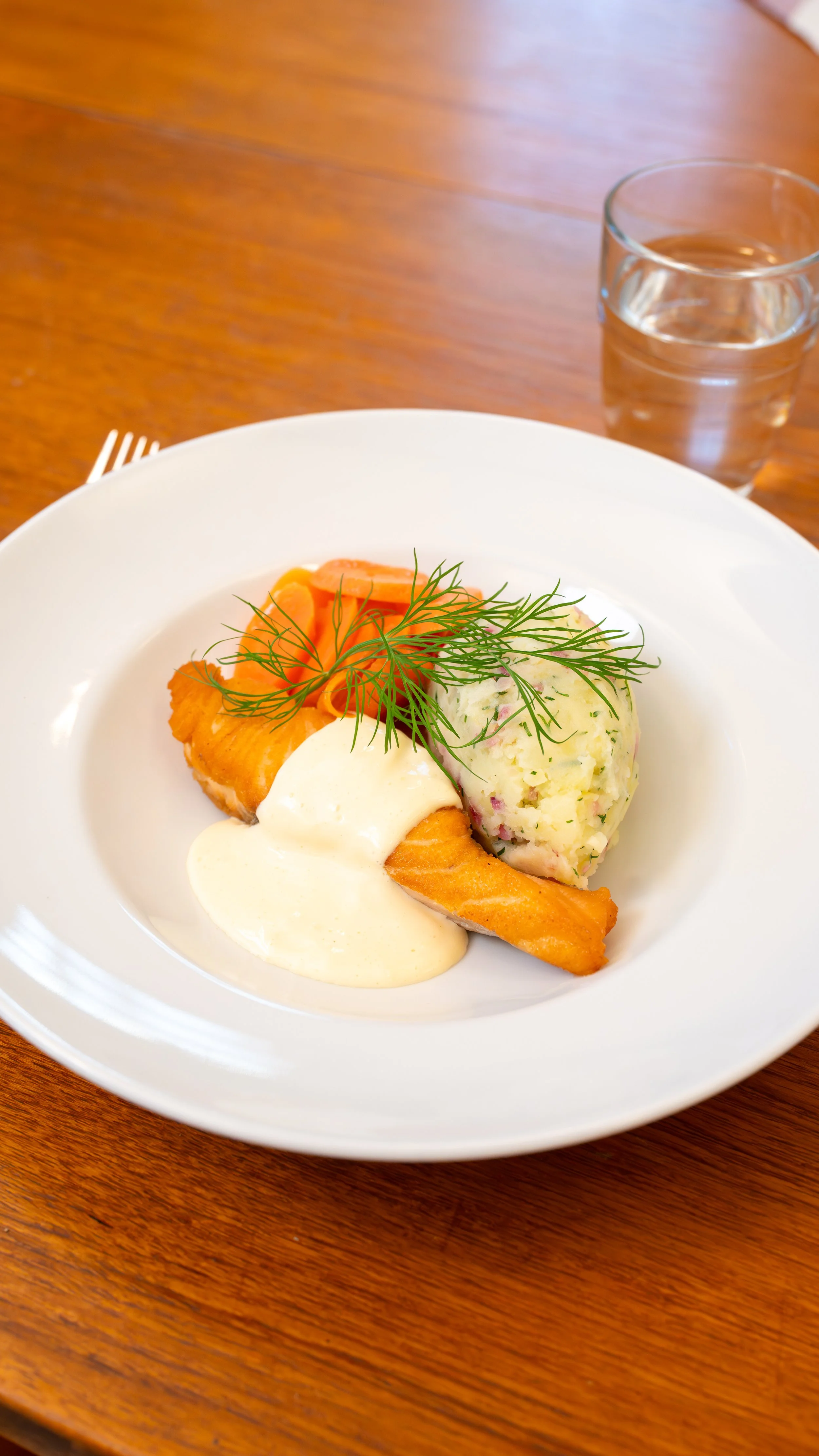 Plate of cooked salmon with dill garnish, mashed potatoes with herbs, and a dollop of creamy sauce, on a white dish. Glass of water on a wooden table.