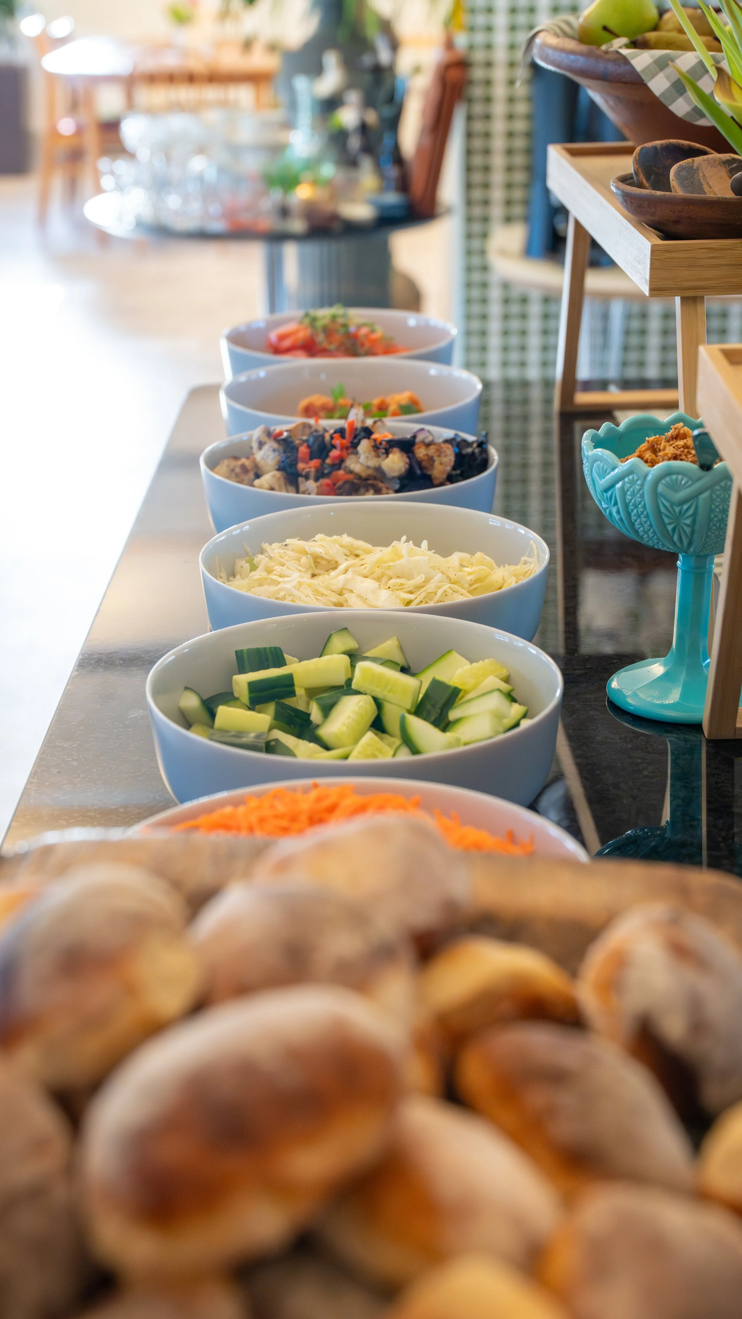 Assorted bowls of chopped vegetables on a table, with bread rolls in the foreground, in a cozy, well-lit indoor setting.