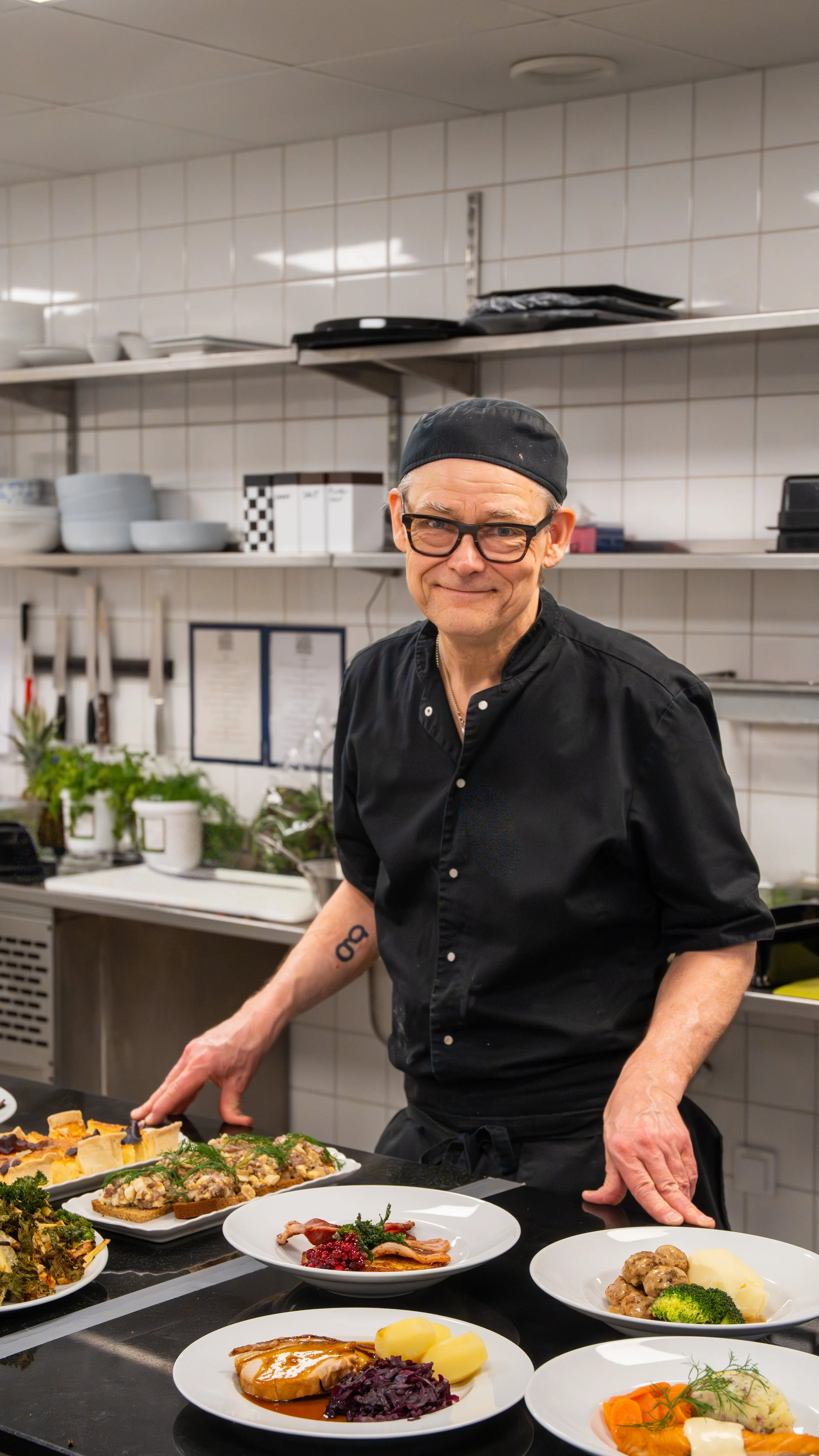 A chef in a black uniform and glasses stands behind a counter with various plated dishes, preparing food in a commercial kitchen.
