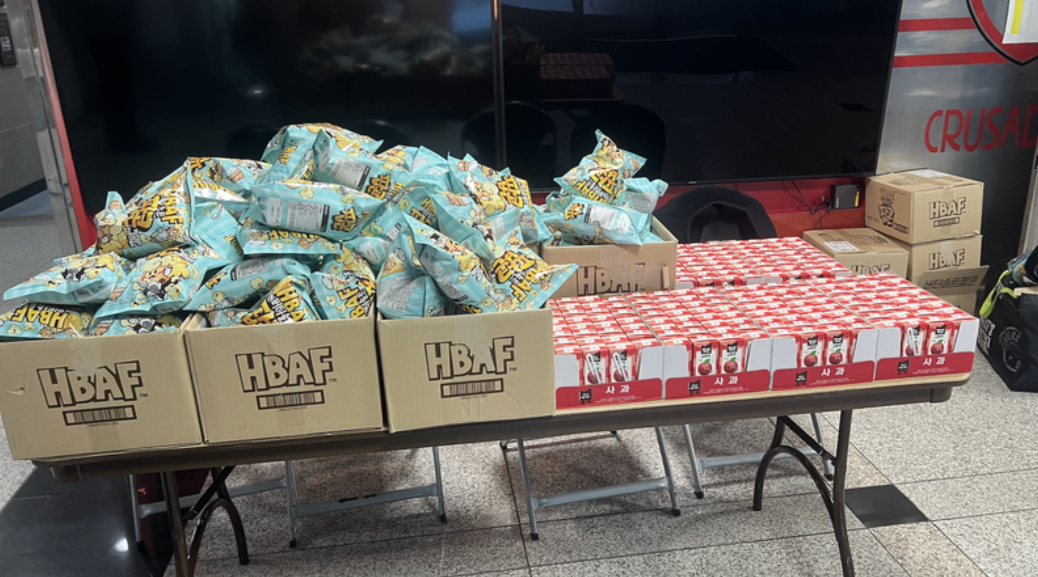 A table with boxes of snacks and drinks. The left side has boxes of 'HBAF' snacks with large bags of chips or popcorn, and the right side has boxes of juice cartons stacked in rows.