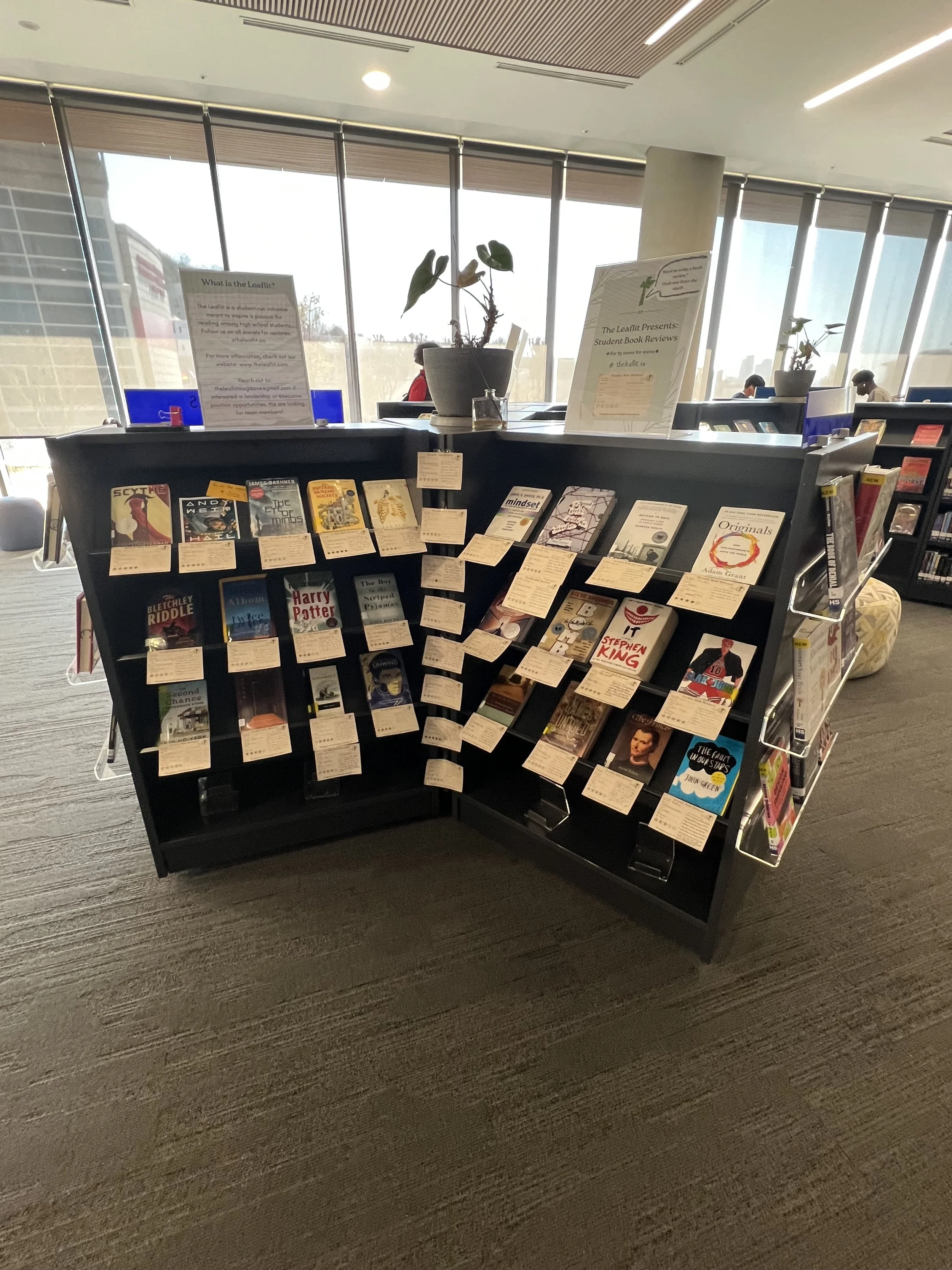 A library bookshelf with books on student book reviews, including titles like 'Harry Potter' and 'It,' with informational signs on top about the reading program.