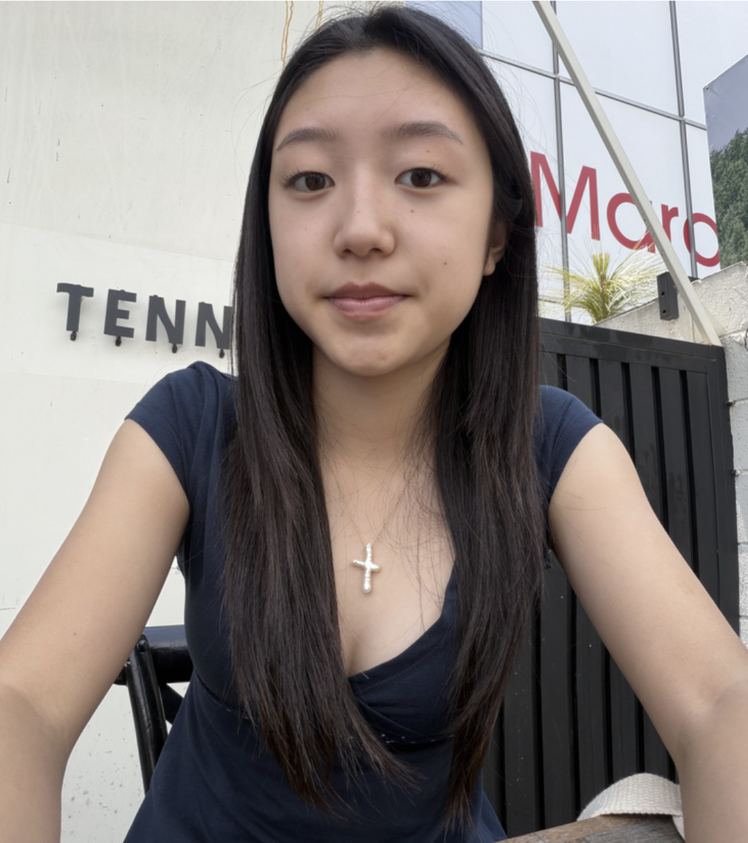 Young woman with long dark hair, wearing a navy blue top and a silver cross necklace, sitting outdoors next to a black fence and a building with signage.