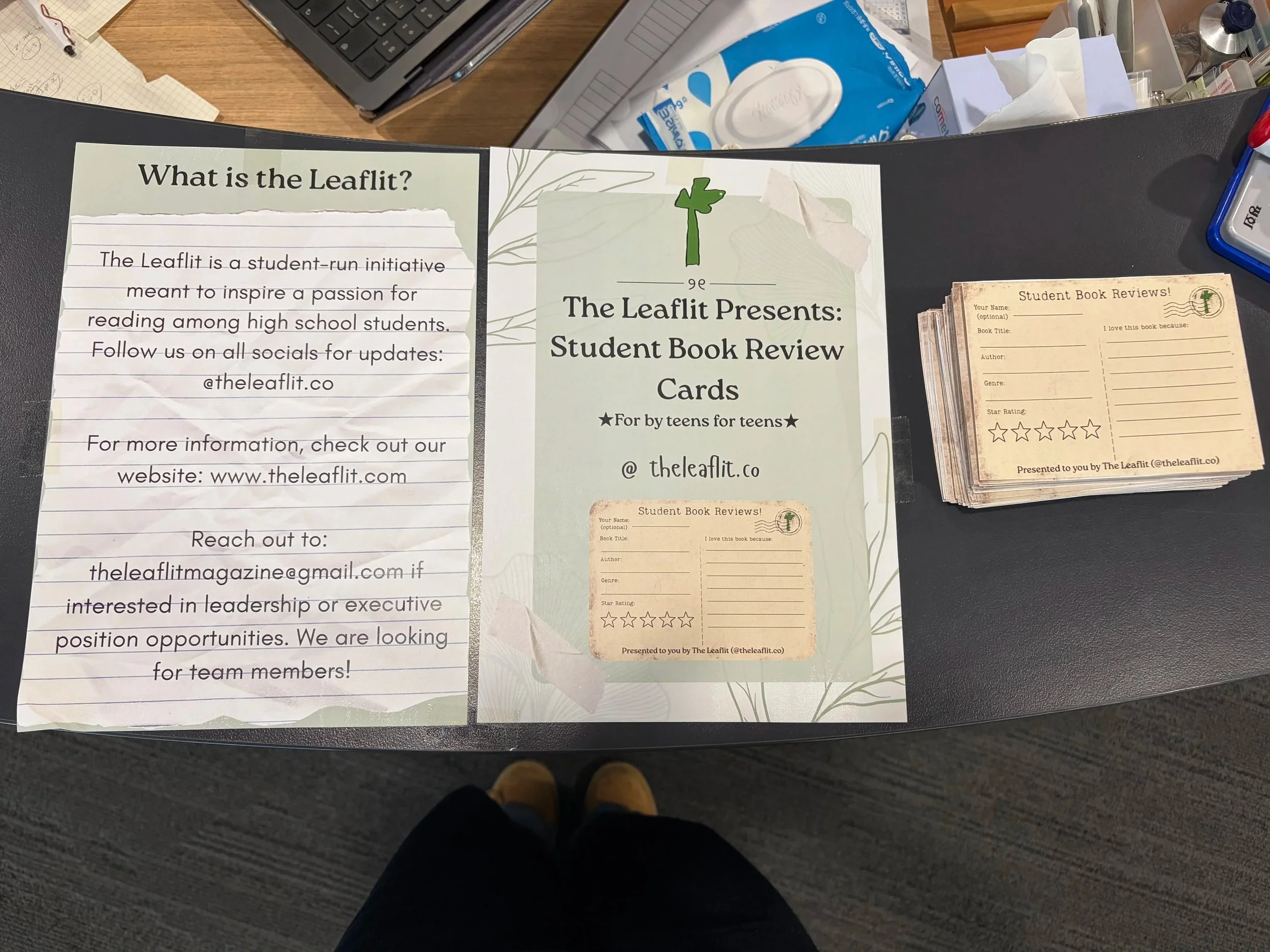 Informational display about the Leaflit student book review initiative, including a description, social media handle '@theleaflit.co', website URL 'www.theleaflit.com', and a call for team members at the bottom left. To the right, there are two stacks of paper review cards for students to write their feedback on books, with sections for title, author, genre, star rating, and reasons for liking the book.