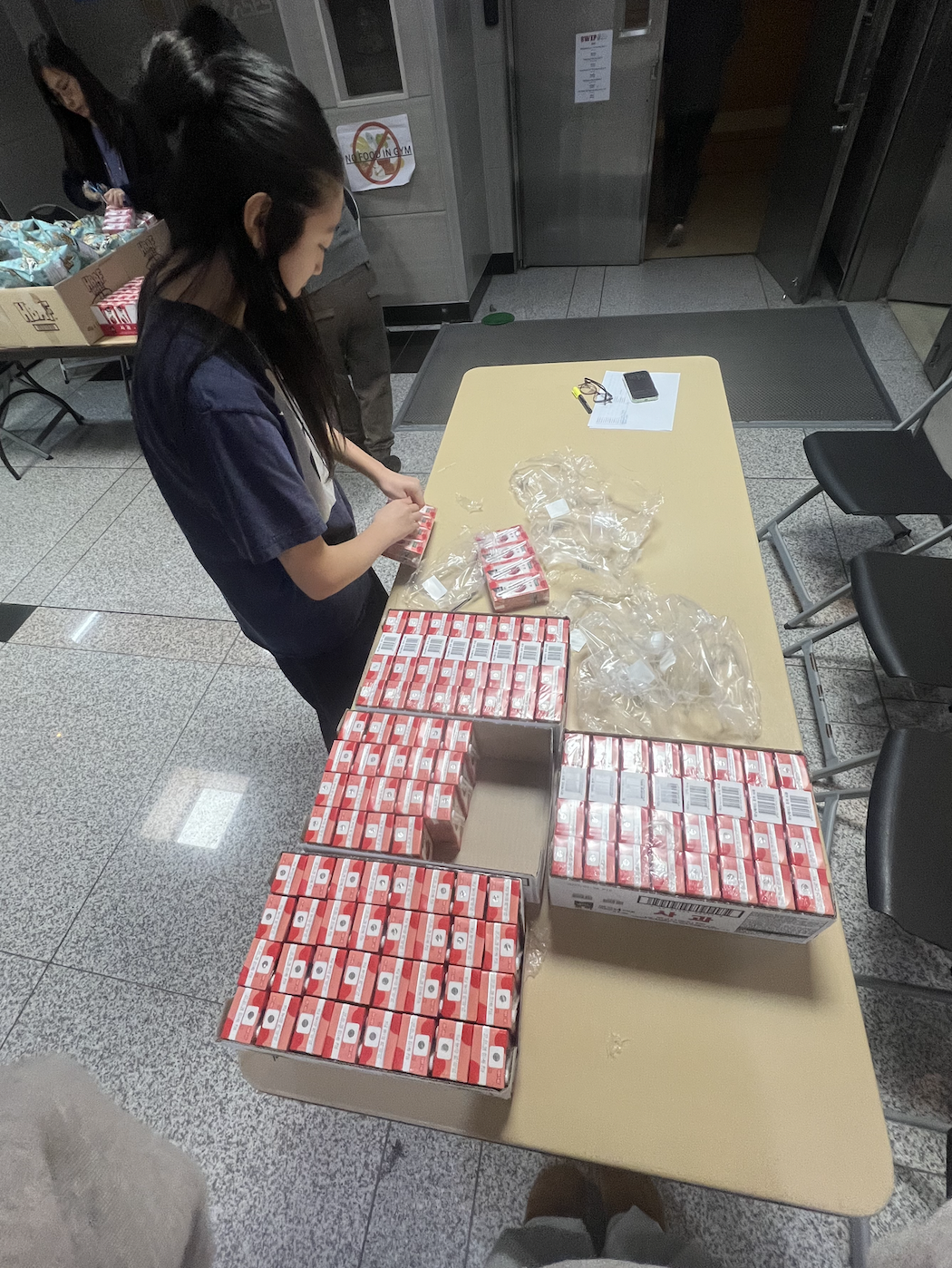 A woman at a table organizing numerous red and white boxes, with a few empty plastic bags and a digital device on the table.