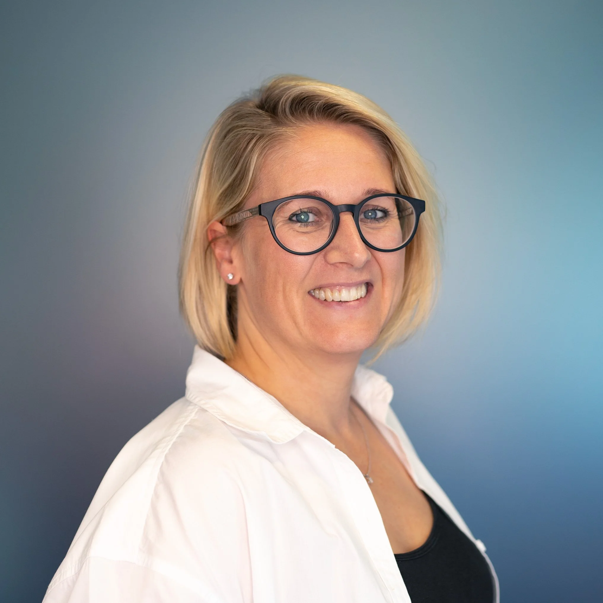 Close-up of a smiling young woman with blonde hair wearing black-rimmed glasses, a white collared shirt, and a black undershirt, against a light blue background.