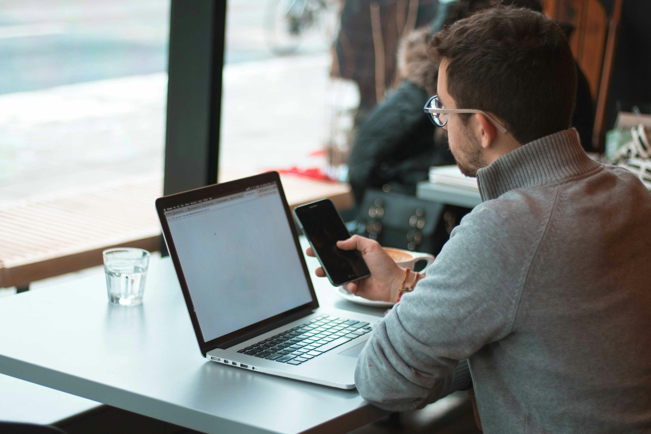 A man with glasses sitting at a table in a cafe, looking at his phone, with a laptop and a glass of water in front of him.