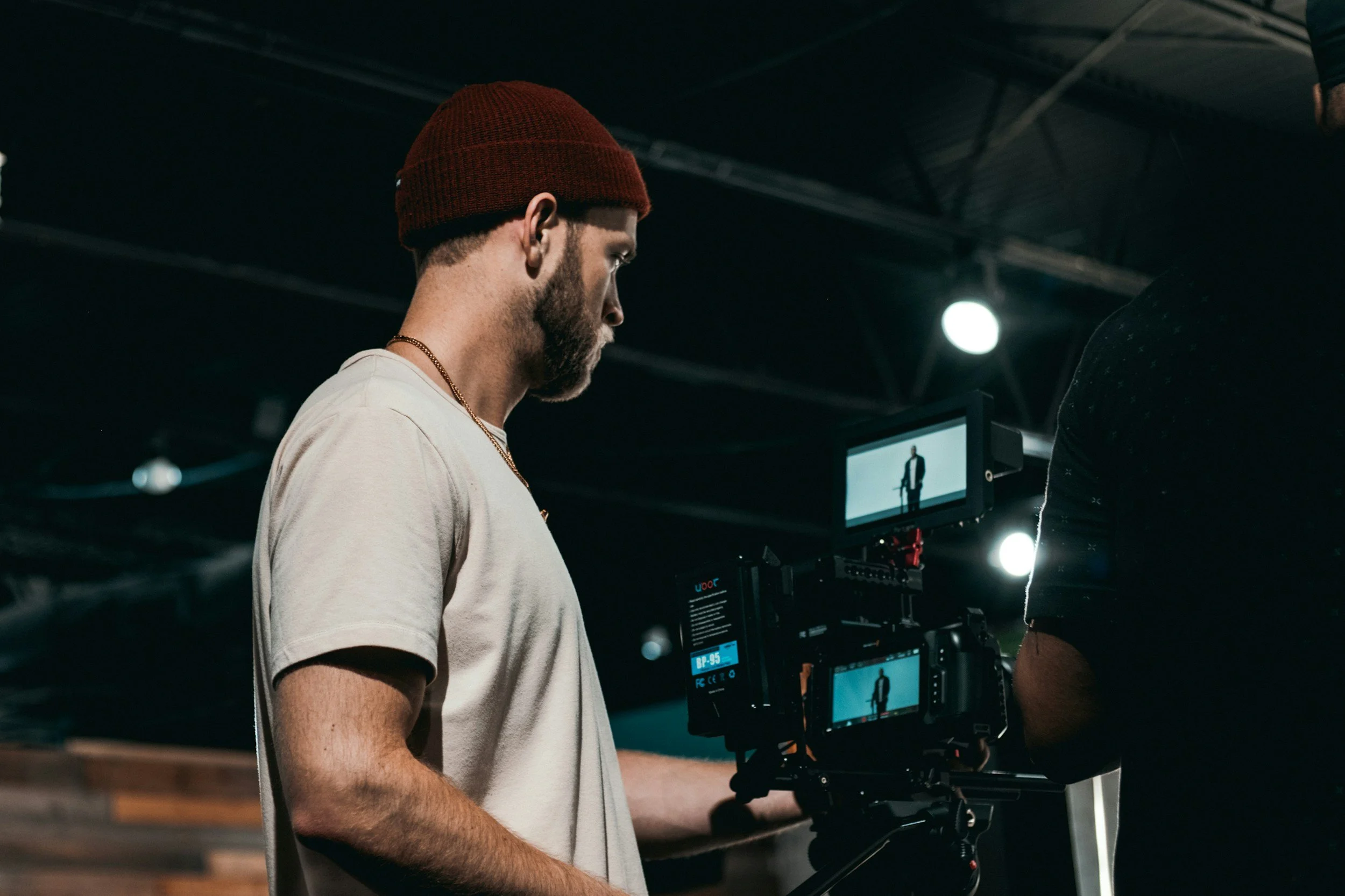 A man with a beard, wearing a red beanie and a white T-shirt, operates a professional camera on a tripod, filming in an indoor studio with dark ceiling and bright lights.